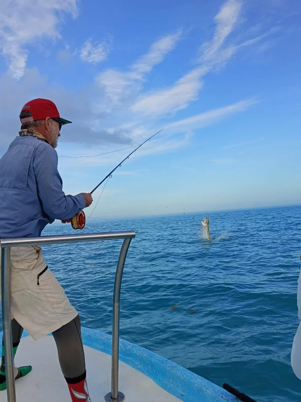 Man fishing on a boat with a rod, catching a fish out of the water, with the ocean and blue sky in the background.