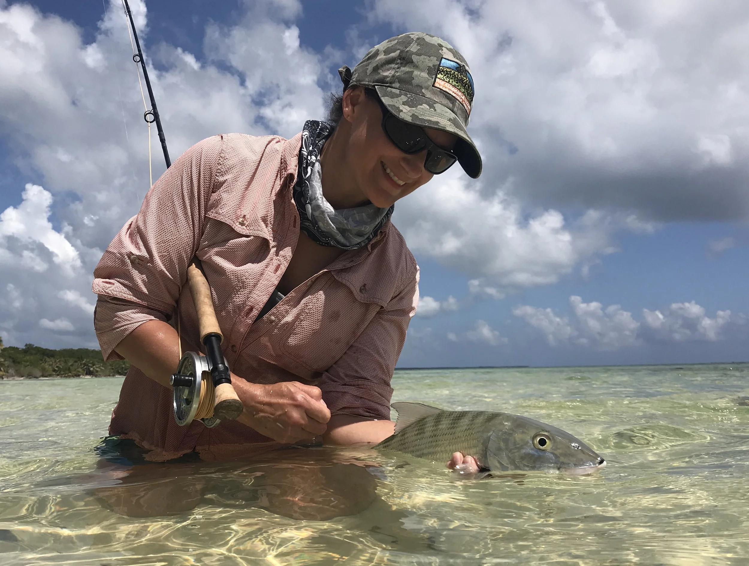 Female angler in pink shirt, sunglasses, and hat kneeling in Turneffe Atoll waters holding a bonefish