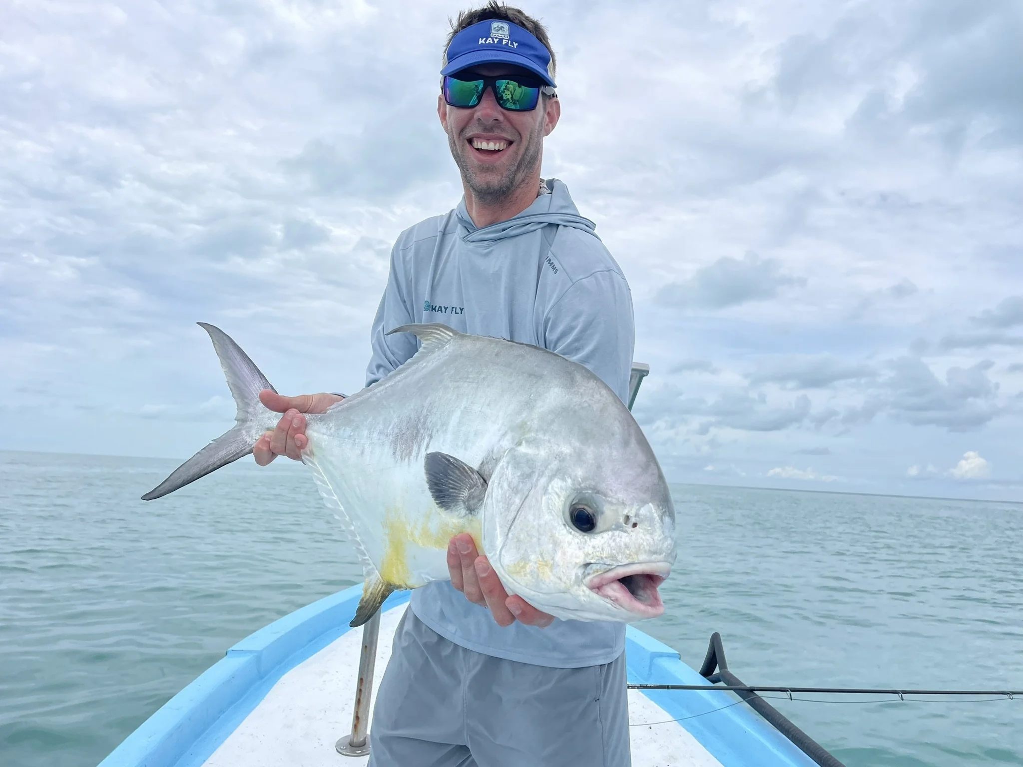 Man on boat holding large fish with open mouth, wearing sunglasses and blue cap, over seascape with cloudy sky.