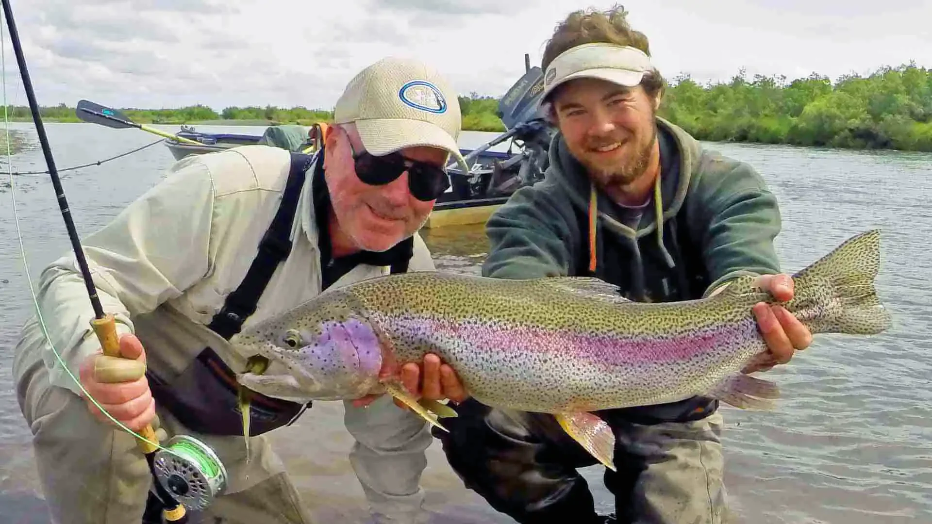 Two happy anglers fly fishing in Alaska with Dave Duncan & Sons, proudly holding a massive, colorful rainbow trout in a freestone river.