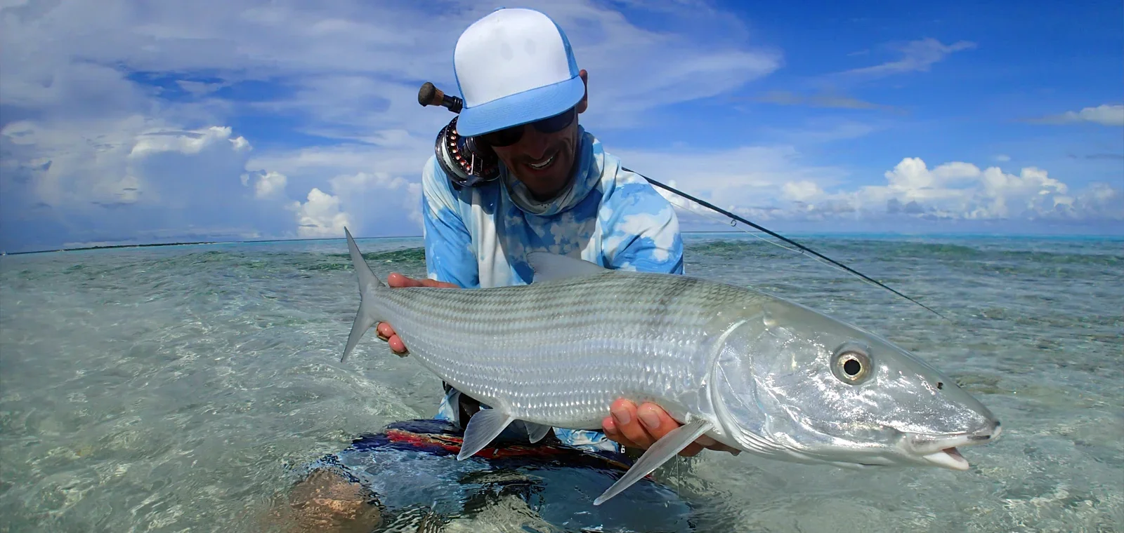 Angler wearing blue and white hat and shirt kneeling in the waters of Christmas Island holding a large bonefish caught while fishing with The Villages