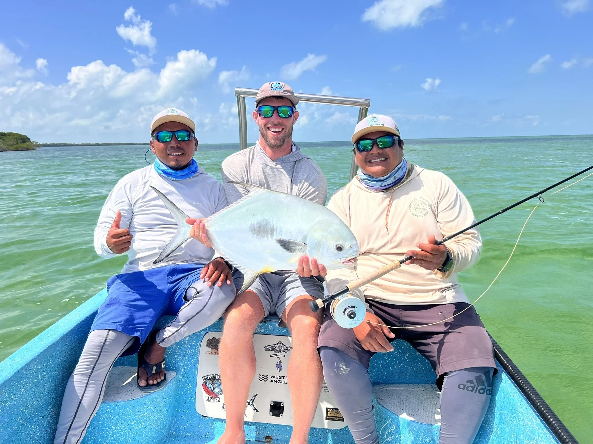 Three men on a boat holding a large fish, with the man in the middle holding the fish in both hands and the two men on the sides smiling and giving thumbs up, in a bright, sunny day on a calm, green water body.