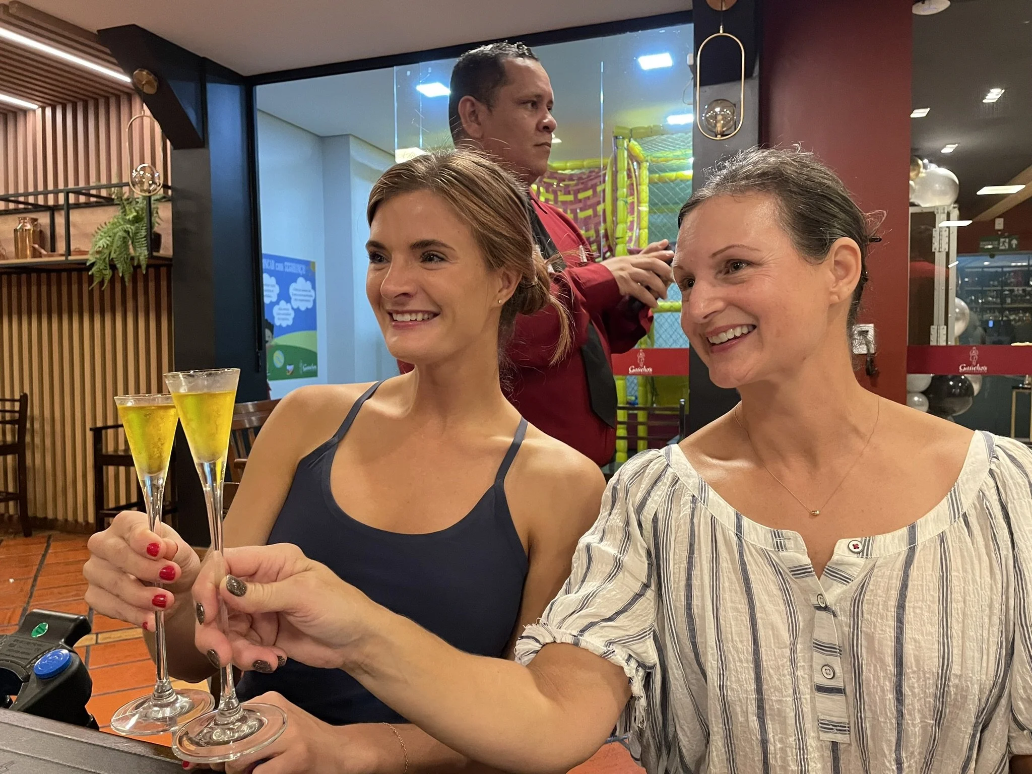 Two women cheerfully clinking glasses of yellow drinks at a restaurant or café in Manaus, Brazil, with a man in a red shirt behind them and wooden decor with colorful indoor play equipment in the background