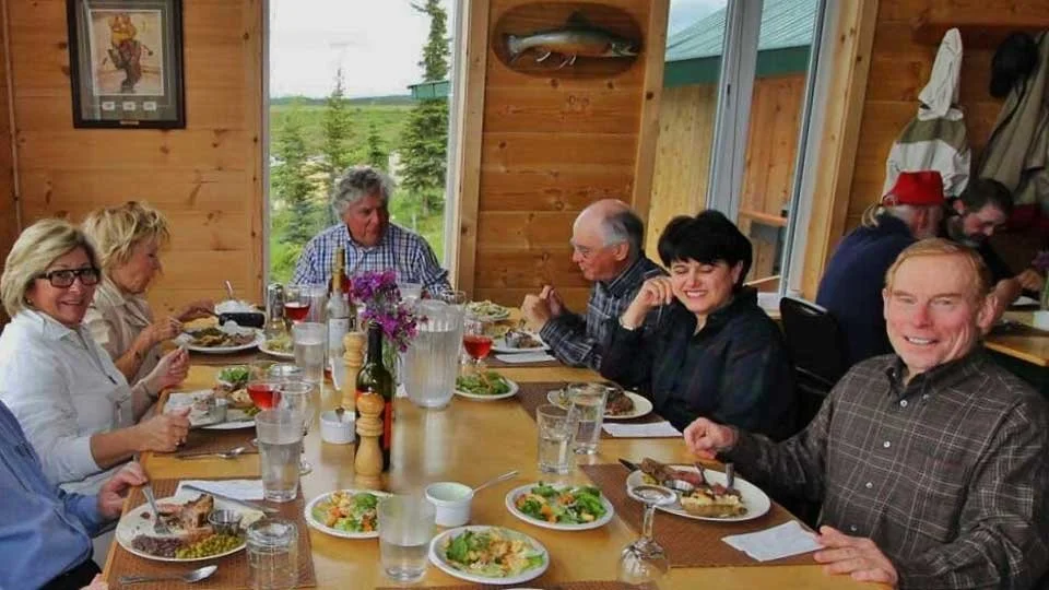 A group of seven people sitting around a wooden dining table inside a cabin, enjoying a meal with plates of food, glasses of water, wine, and a salad. The cabin has wood-paneled walls and a window overlooking a green landscape.