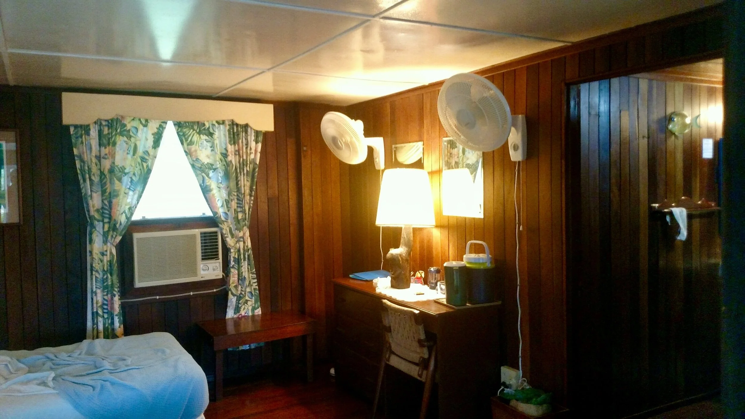 Interior of a rustic guest room at Belize River Lodge with wood paneling, bed, window, AC, dresser, and fans.
