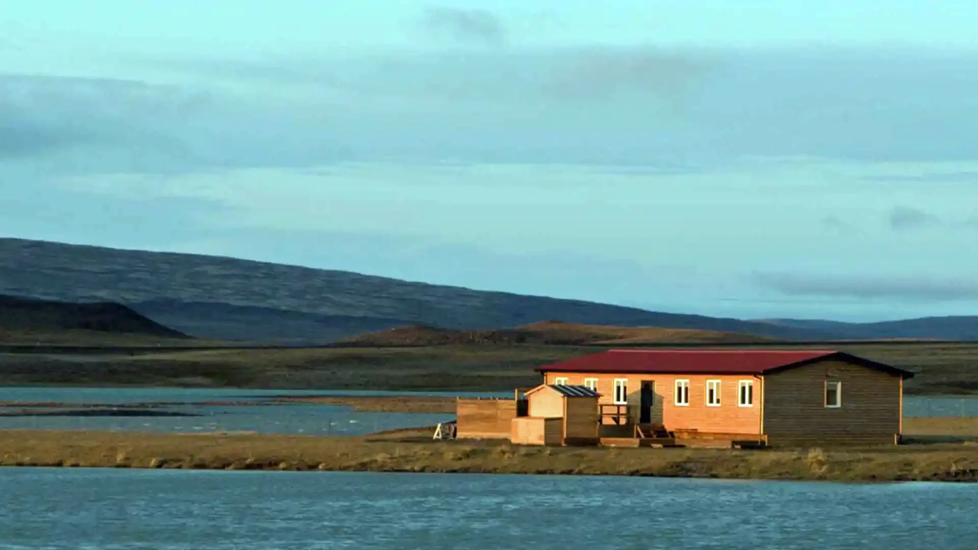 Remote wooden Highland fishing lodge with red roof by a tranquil lake in the rugged Icelandic landscape, ideal for trout fishing.