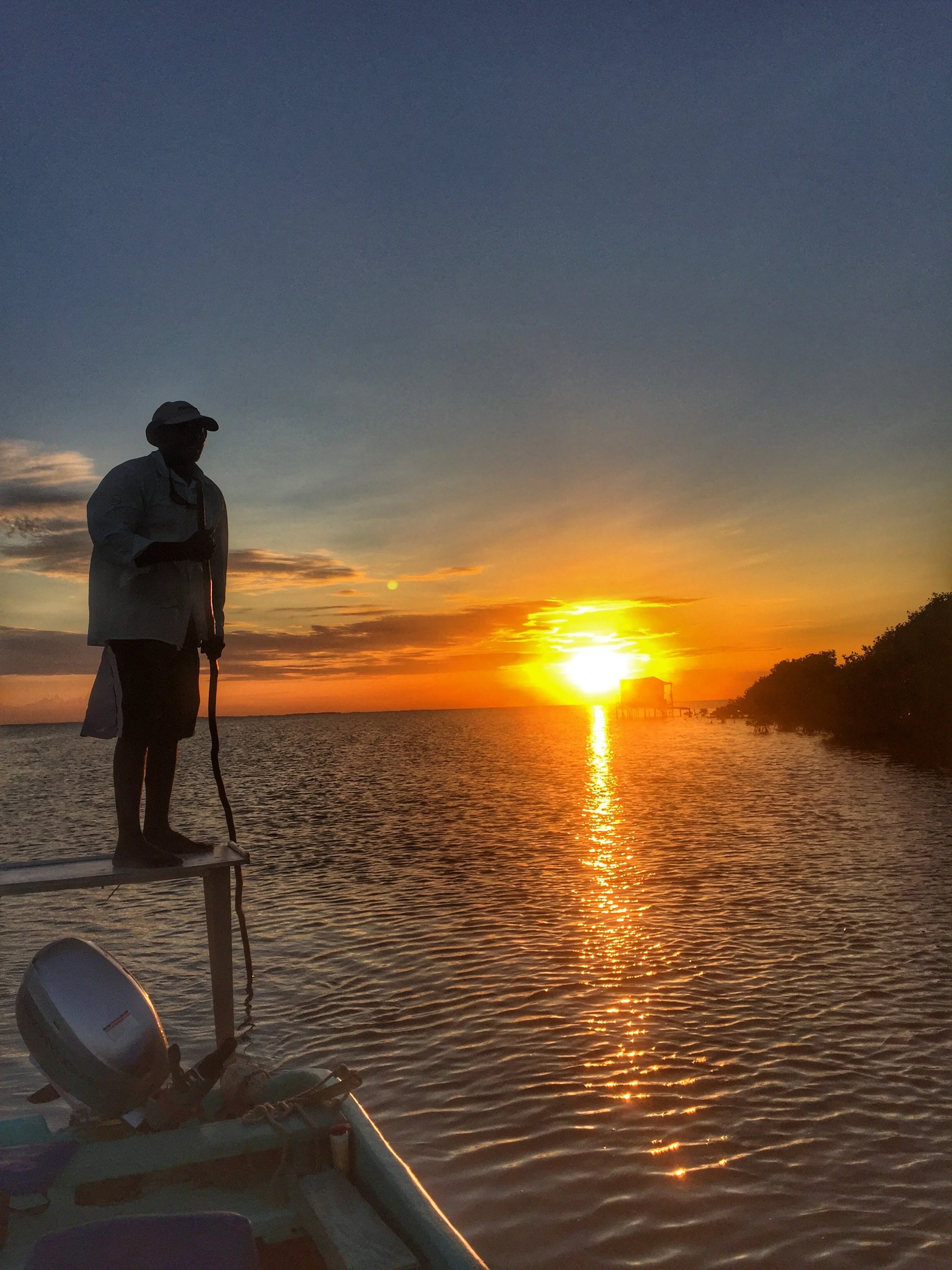 Silhouette of a man standing on a boat during a vibrant sunset over water, with trees on the right side.