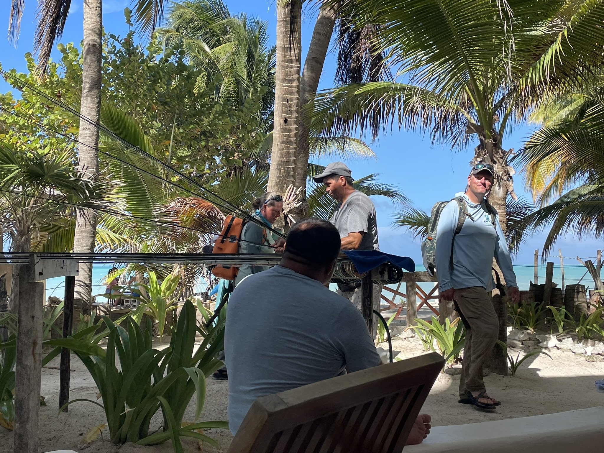A group of people on a tropical beach with palm trees, some are sitting and others standing, engaged in conversation and looking at each other. The scene includes a person sitting with their back to the camera, and others wearing casual outdoor cloth