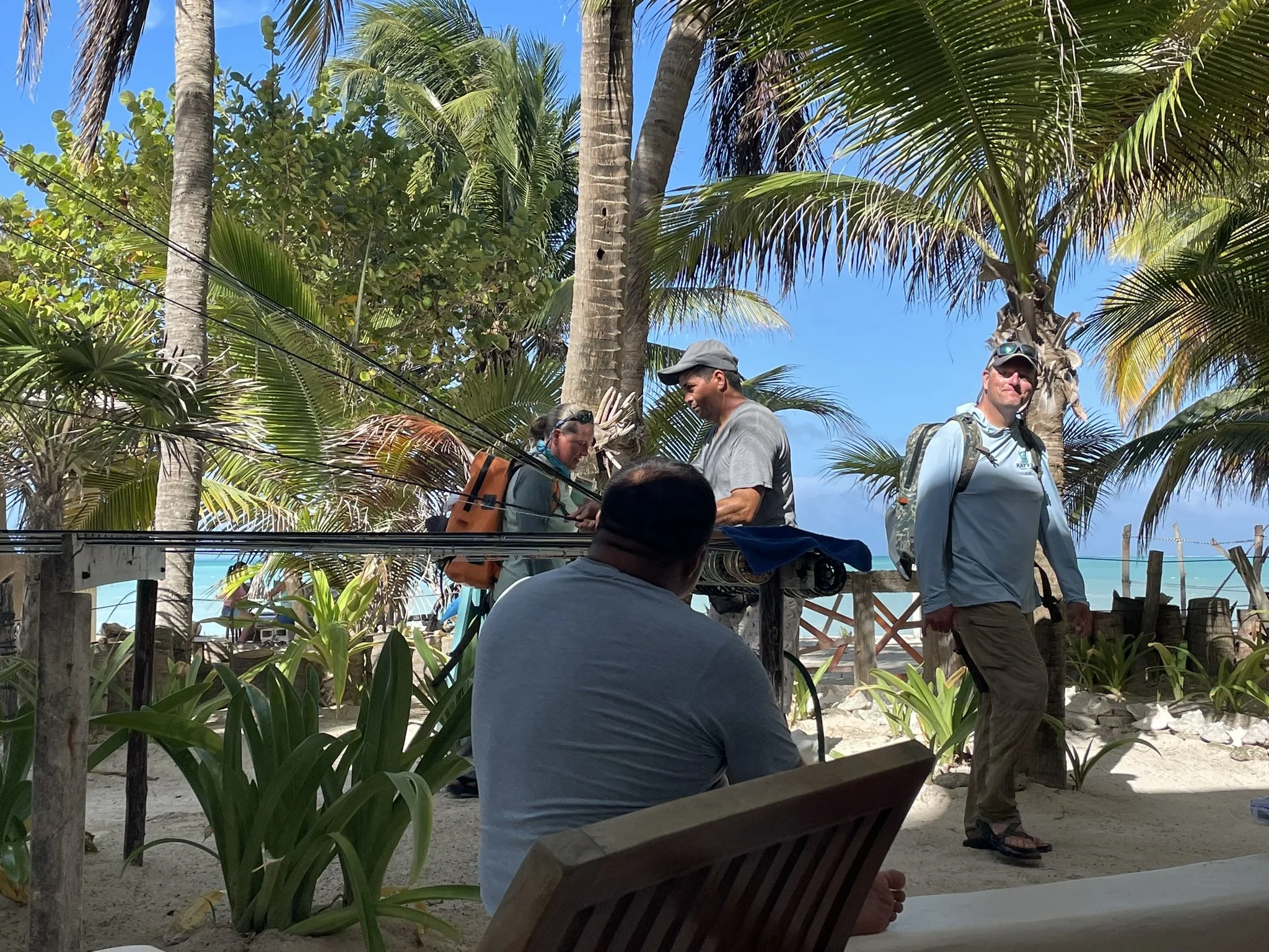 Group of people returning from a day of fishing to Kay Fly Lodge on a tropical beach with palm trees, some sitting and others standing, chatting and looking at each other, with blue sky, turquoise ocean, and lush greenery in the background.