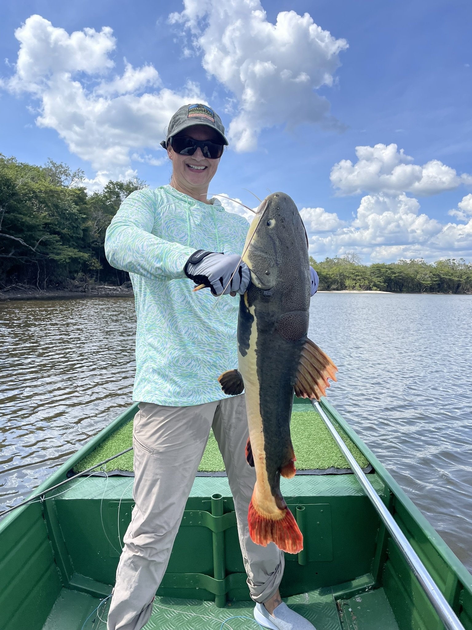 A woman smiling on a boat holding a large catfish she caught, with trees and blue sky with clouds in the background.