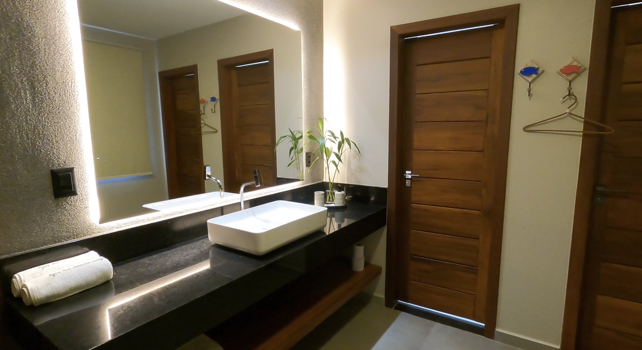 Bathroom with wooden doors, a large mirror, a black countertop with a white rectangular sink, a towel, a plant, and decorative fish hooks.