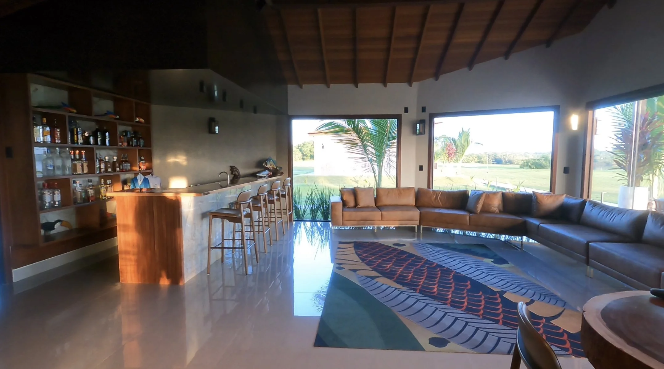 Interior of a modern living room with large windows overlooking a golf course, featuring a brown leather sectional sofa, a patterned area rug, and a bar area with stools.