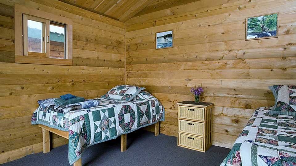 A cozy bedroom with wooden paneled walls, featuring two beds with patterned quilts, a small wicker nightstand with flowers, a window showing an outdoor view, and three framed photos on the wall.