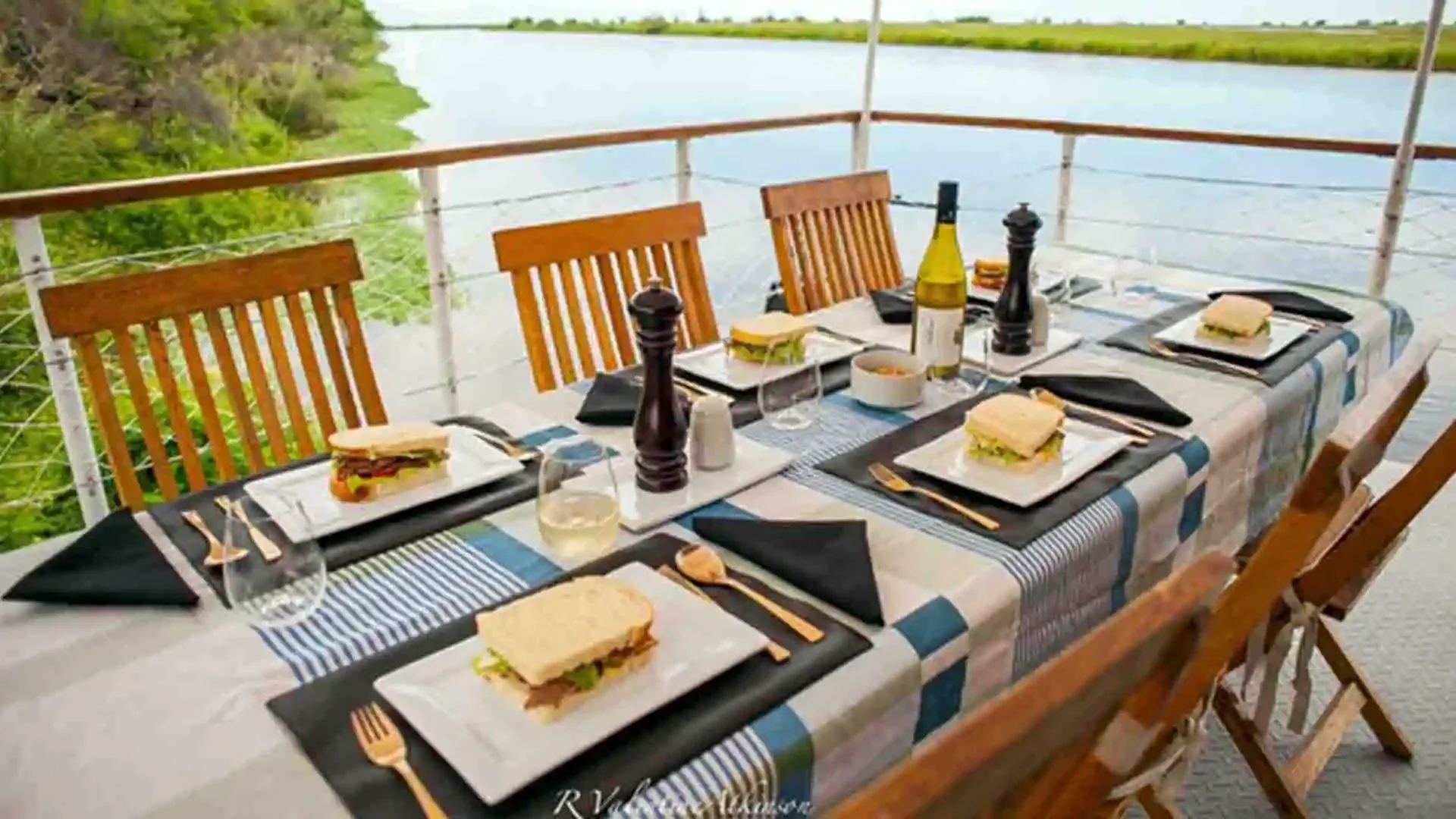 An outdoor dining setup on a boat deck overlooking a river. The table is set with sandwiches, black napkins, wine glasses, bottles of olive oil, and salt and pepper shakers. Wooden chairs surround the table.