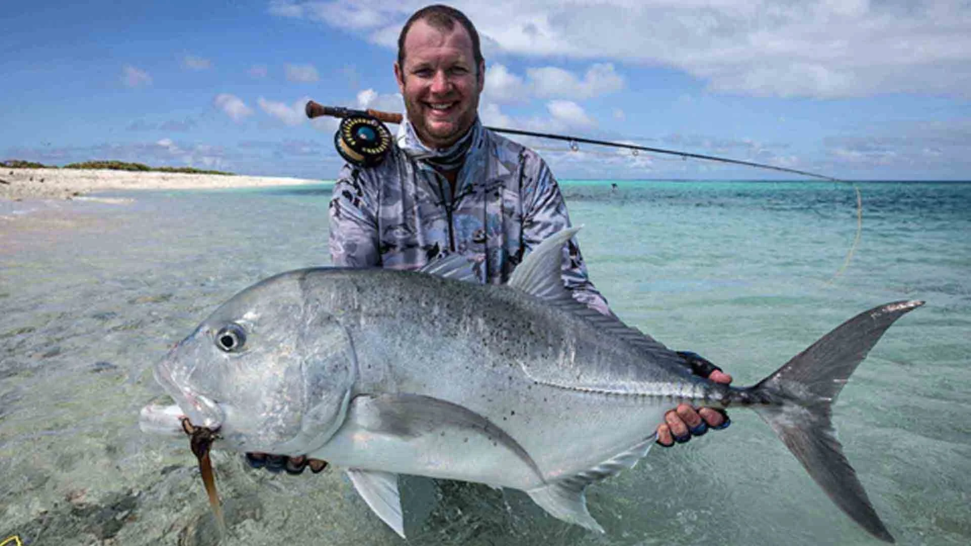 Angler standing in the crystal-clear waters of Farquhar Atoll, Seychelles, holding a Giant Trevally with a fly rod over his shoulder during a saltwater fly-fishing trip.