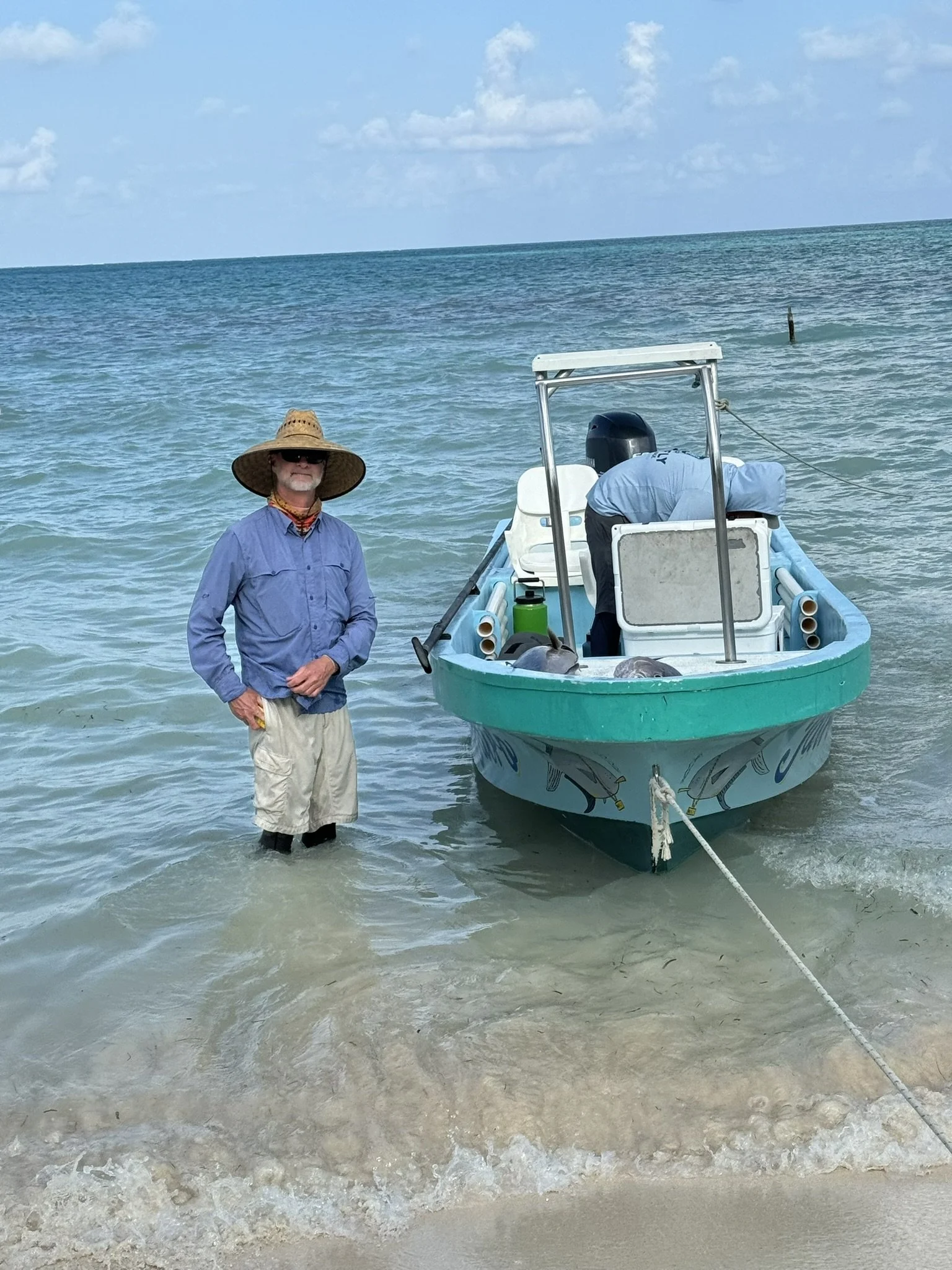 Man wearing a wide-brimmed straw hat, sunglasses, blue shirt, and shorts standing in shallow water next to a Kay Fly Lodge fishing boat at the shoreline of Ascension Bay.
