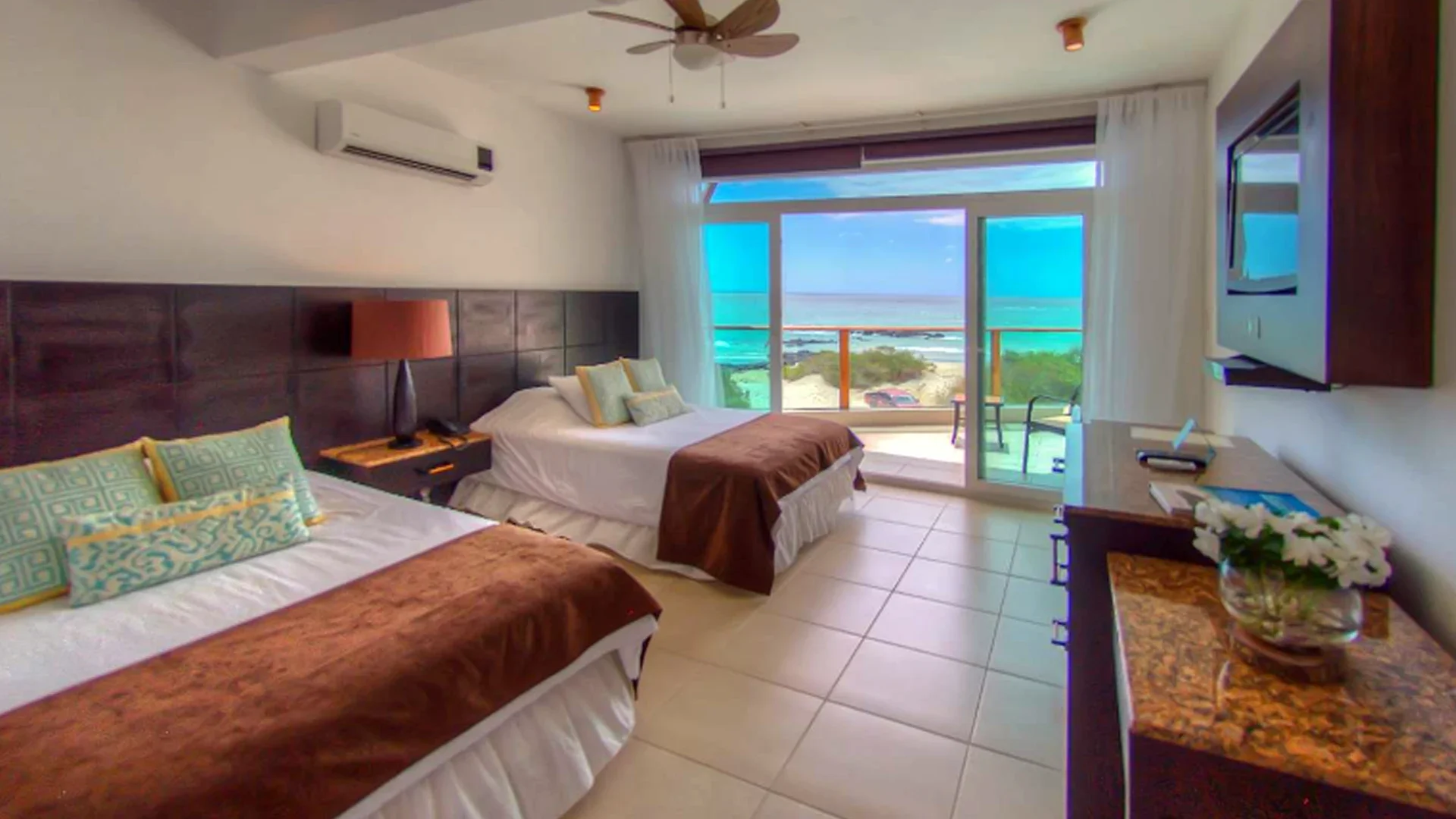 Hotel room with two beds, large window showing ocean view, balcony with chairs, and wall-mounted TV.