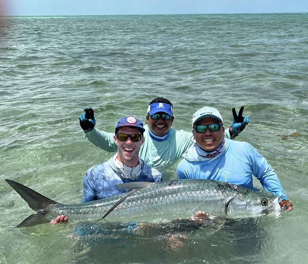 Three smiling anglers standing in the clear waters of Ascension Bay holding a freshly caught tarpon while fishing with Kāy Fly Lodge
