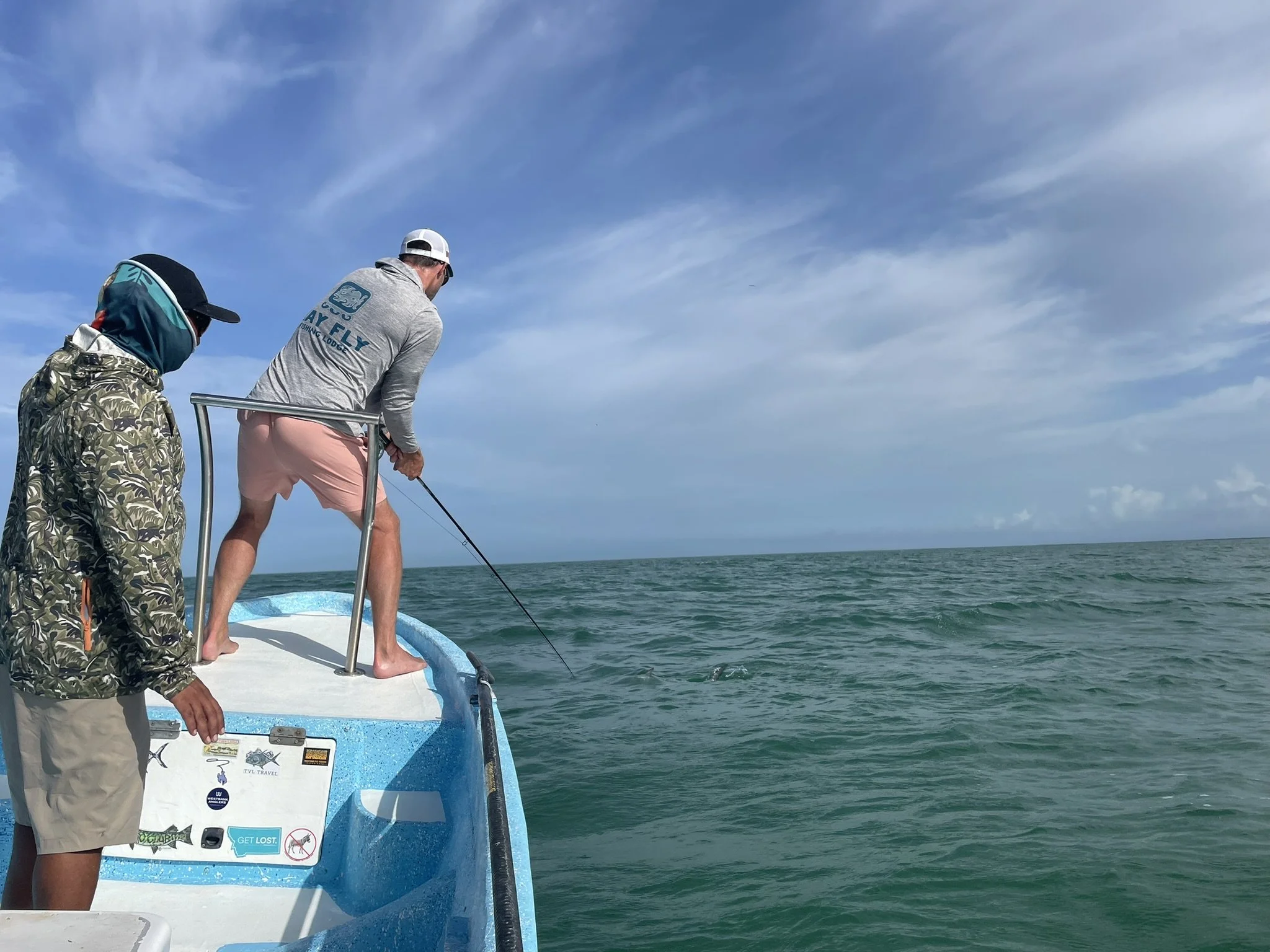 Two men fishing from a boat on a clear day in Ascension Bay, with one fighting a tarpon while the Kay Fly Lodge guide watches, and blue sky and ocean in the background.