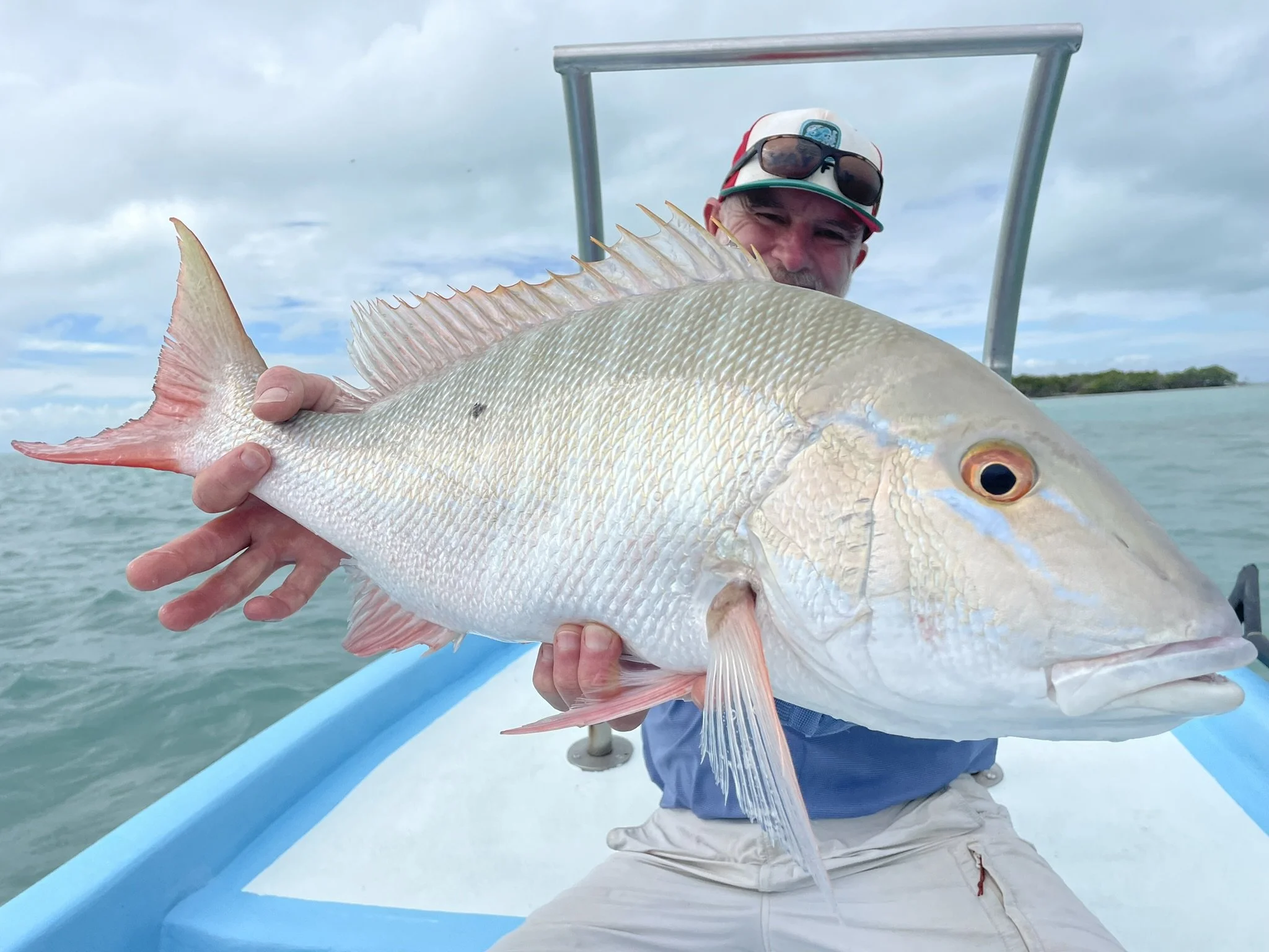 A man in a boat holding a large fish with a pale body, pink fins, and a prominent eye, smiling at the camera against a cloudy sky and water background.
