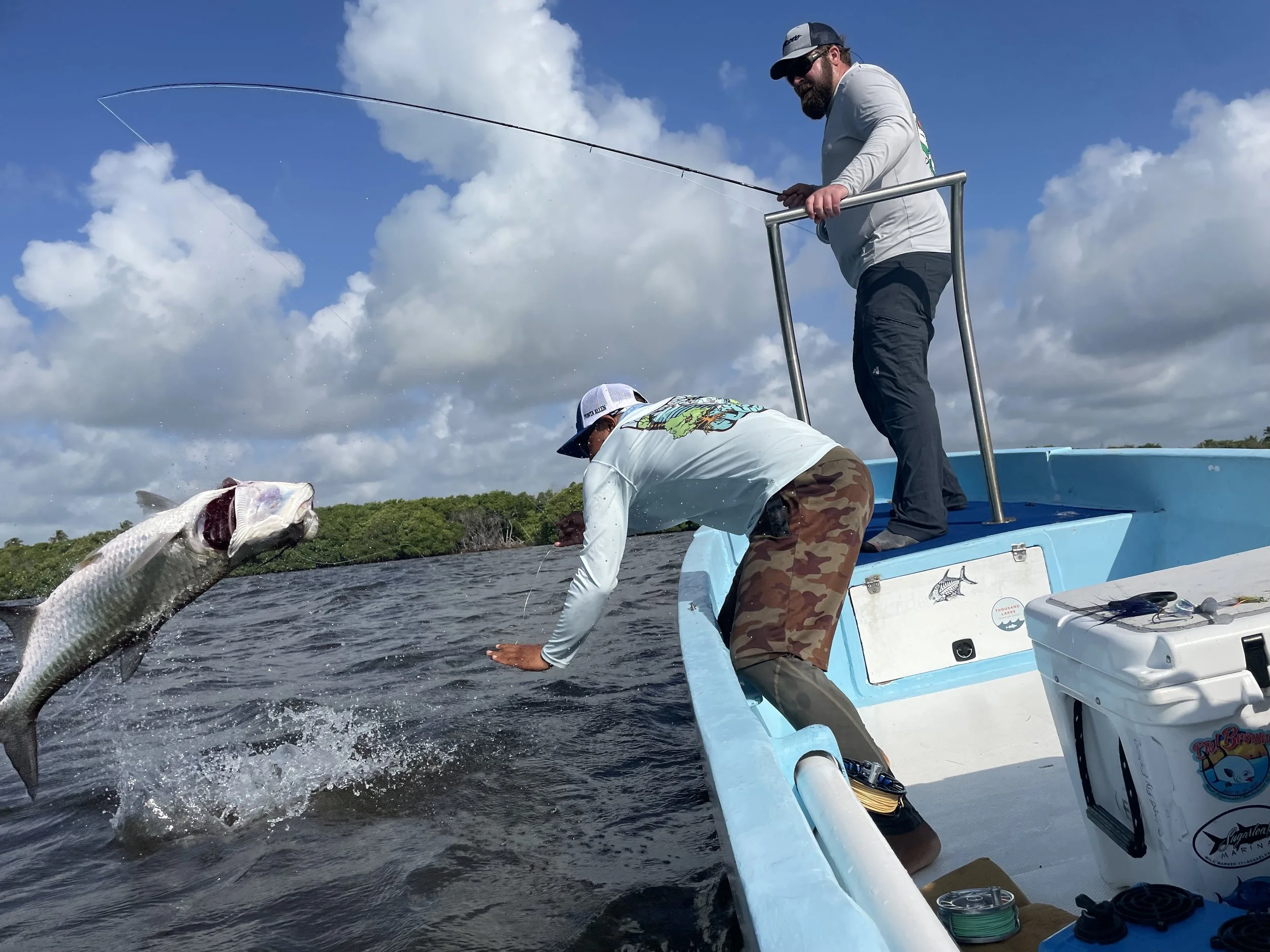 Two men on a boat landing a tarpon, with one holding a fishing rod and the guide from Kay Fly Lodge leaning over the side under a partly cloudy sky.