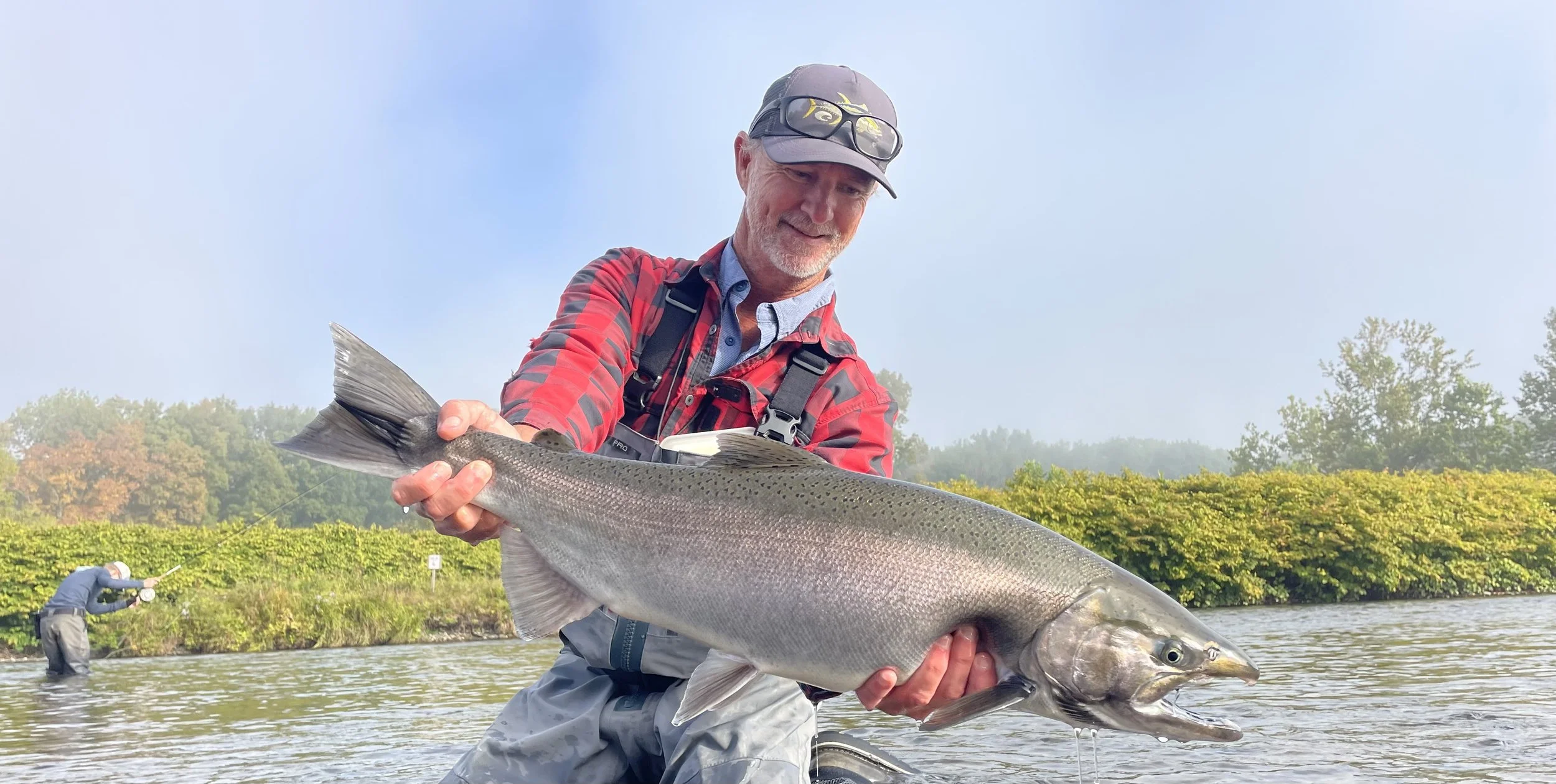 A man holding a large fish with a smile, standing in a river with another person fishing in the background and trees on the riverbank.