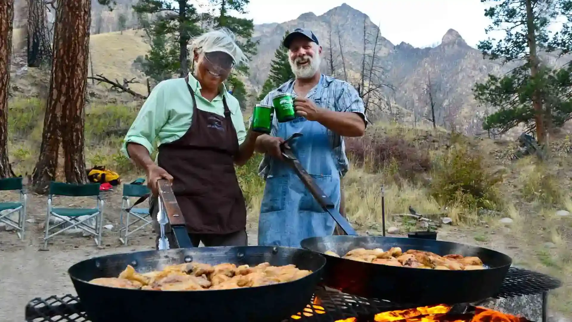 Two men cook chicken over a campfire on the Middle Fork of the Salmon River, enjoying a delicious wilderness camp meal.