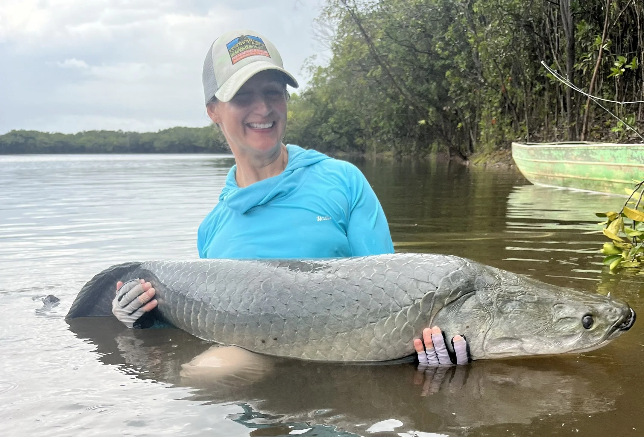 Woman in a blue long-sleeve shirt and a cap smiling while holding a large fish in shallow water, with a boat and trees in the background.