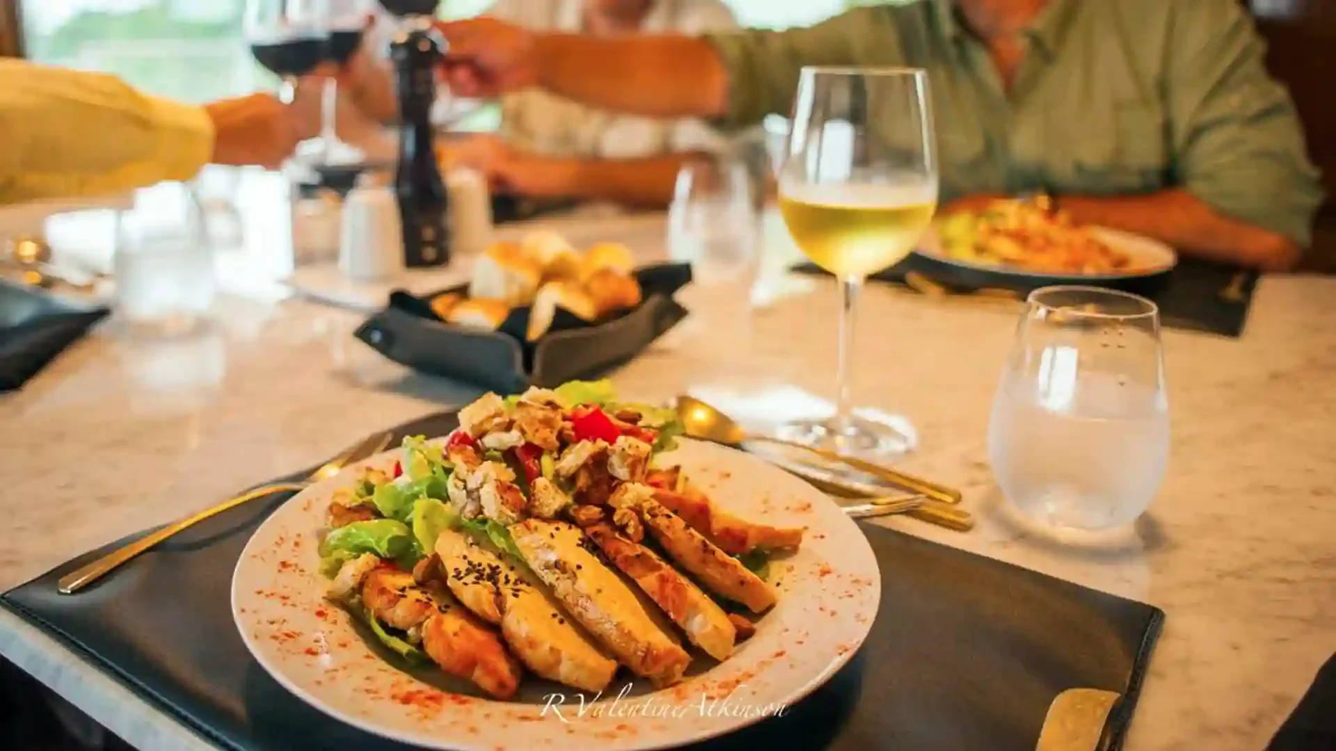 A dinner table set with a plate of grilled chicken and salad, a glass of white wine, a glass of water, and a basket of bread. There are people in the background, one in a green shirt and another in a yellow shirt, reaching for food.