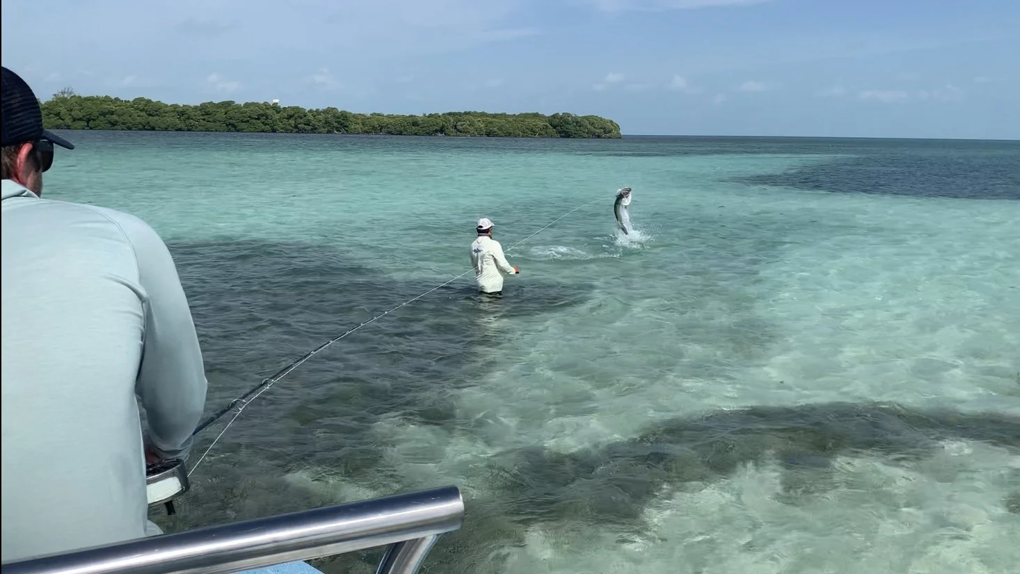 Two men fishing in shallow clear water, one is reeling in a fish that is leaping out of the water, with a tropical island in the background.