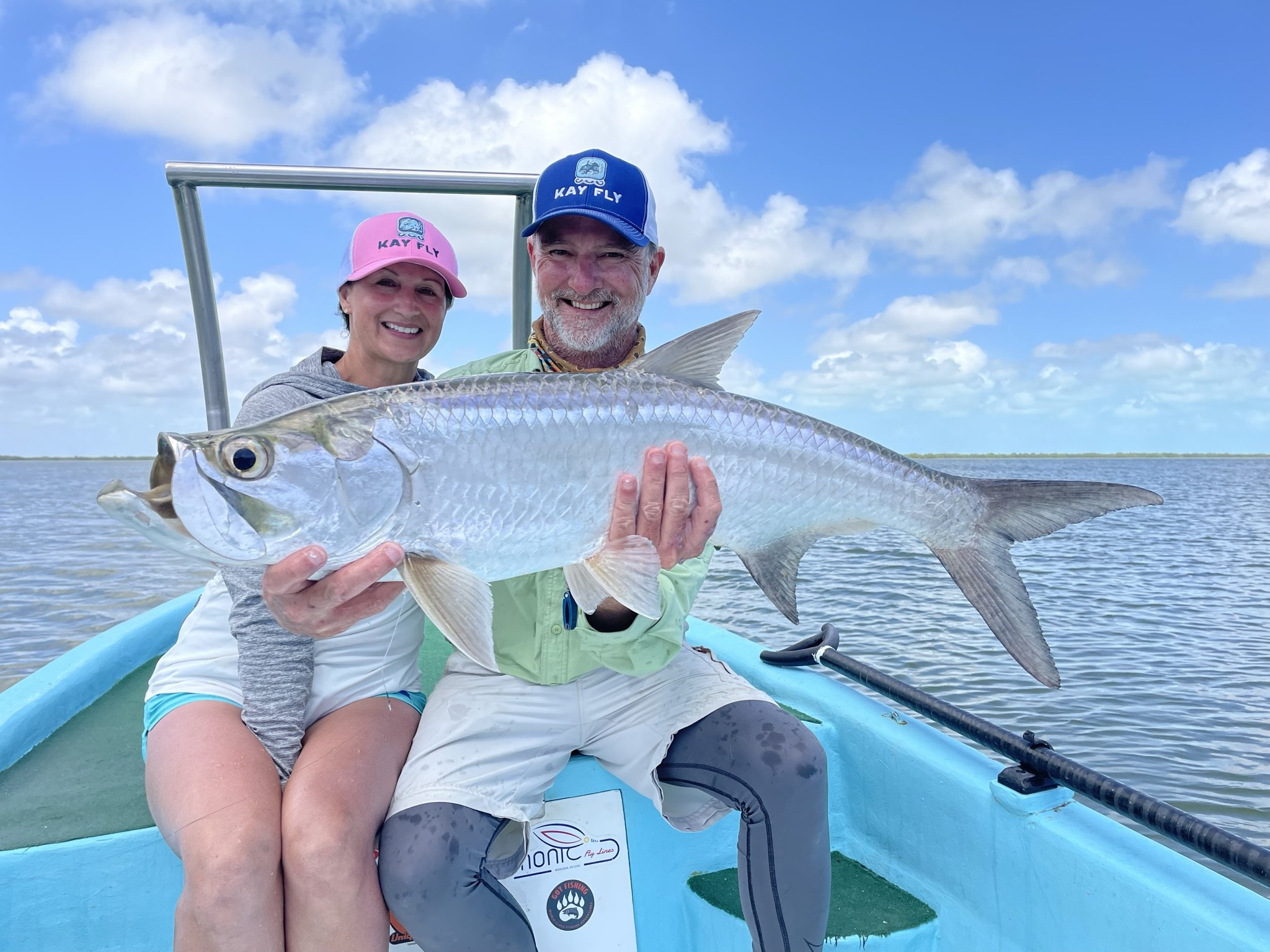 Two people, a man and a woman, on a boat holding a large fish they caught, with water and sky in the background.