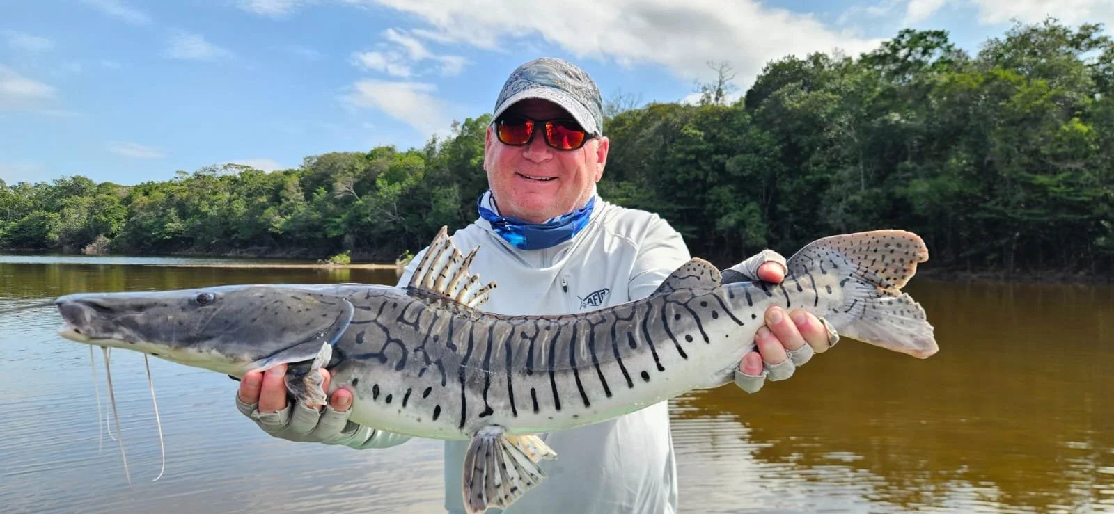 A man wearing sunglasses, a hat, and a long-sleeve shirt holding a large fish by a river with trees in the background.