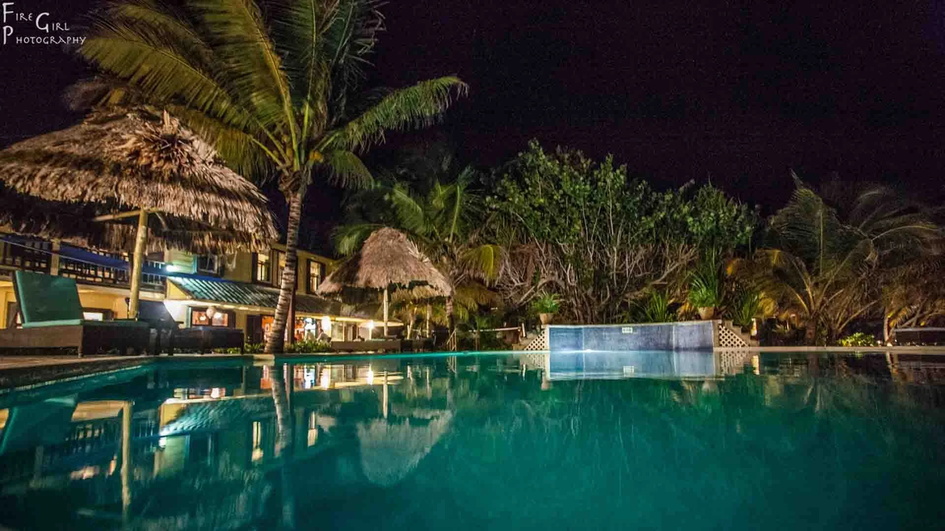 Nighttime view of the illuminated swimming pool and main lodge at El Pescador Lodge, Ambergris Caye, Belize, surrounded by tropical palm trees.