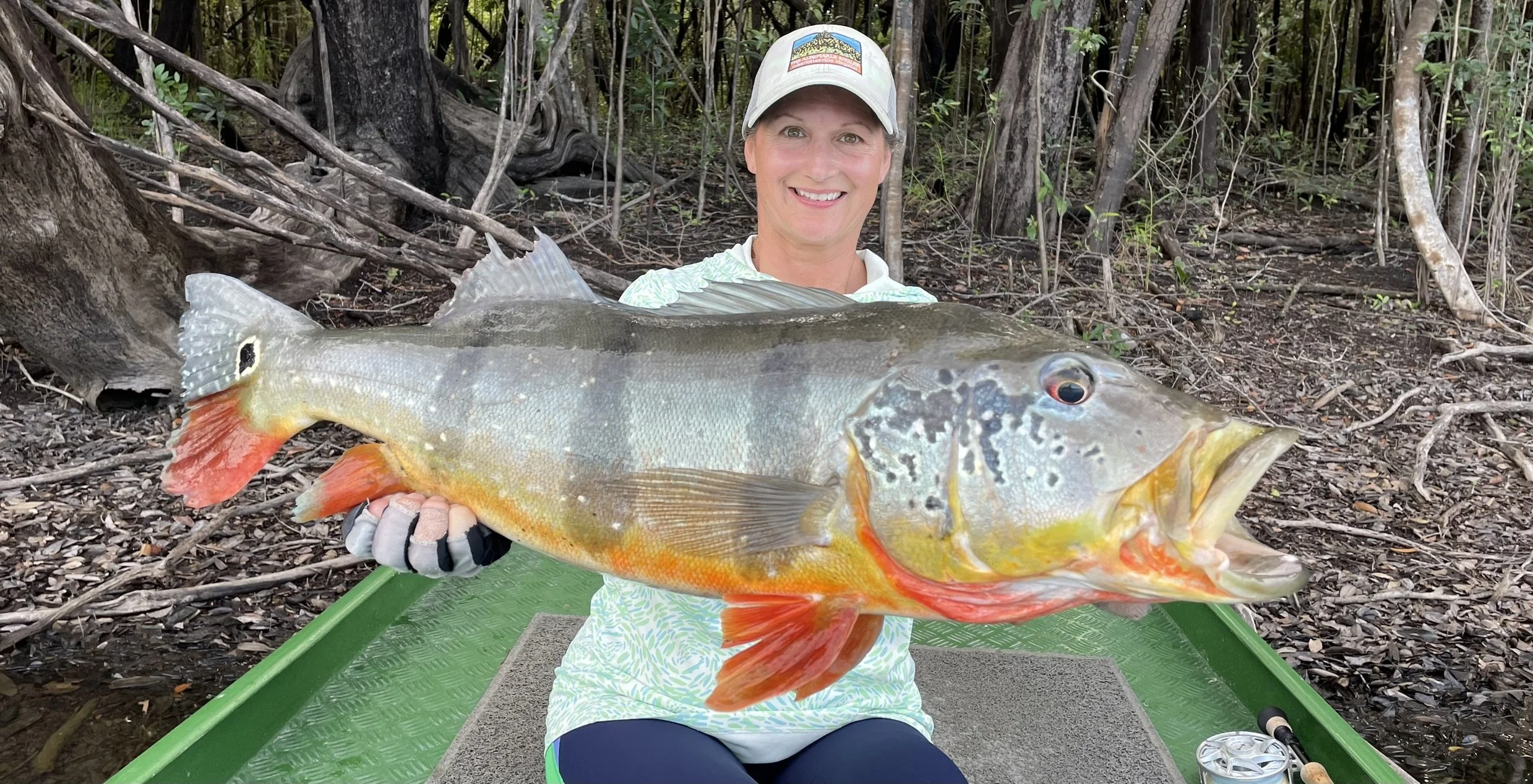 Woman smiling while holding a large peacock bass at Agua Boa Amazon Lodge in a forest setting with trees and roots in the background.