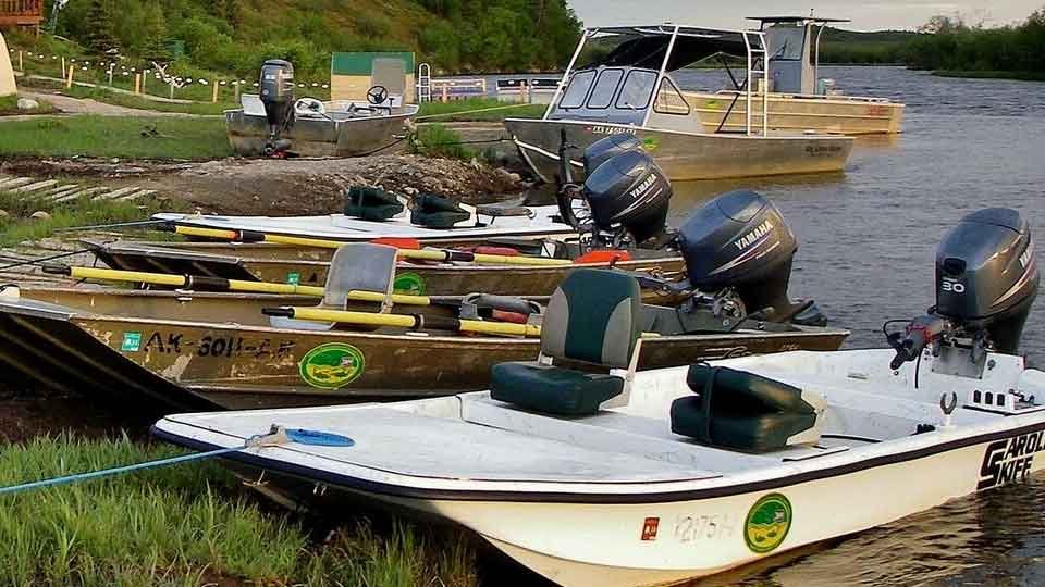 Several boats docked at the water's edge, equipped with outboard motors, with paddles and gear inside the boats, and a larger boat in the background on the water.