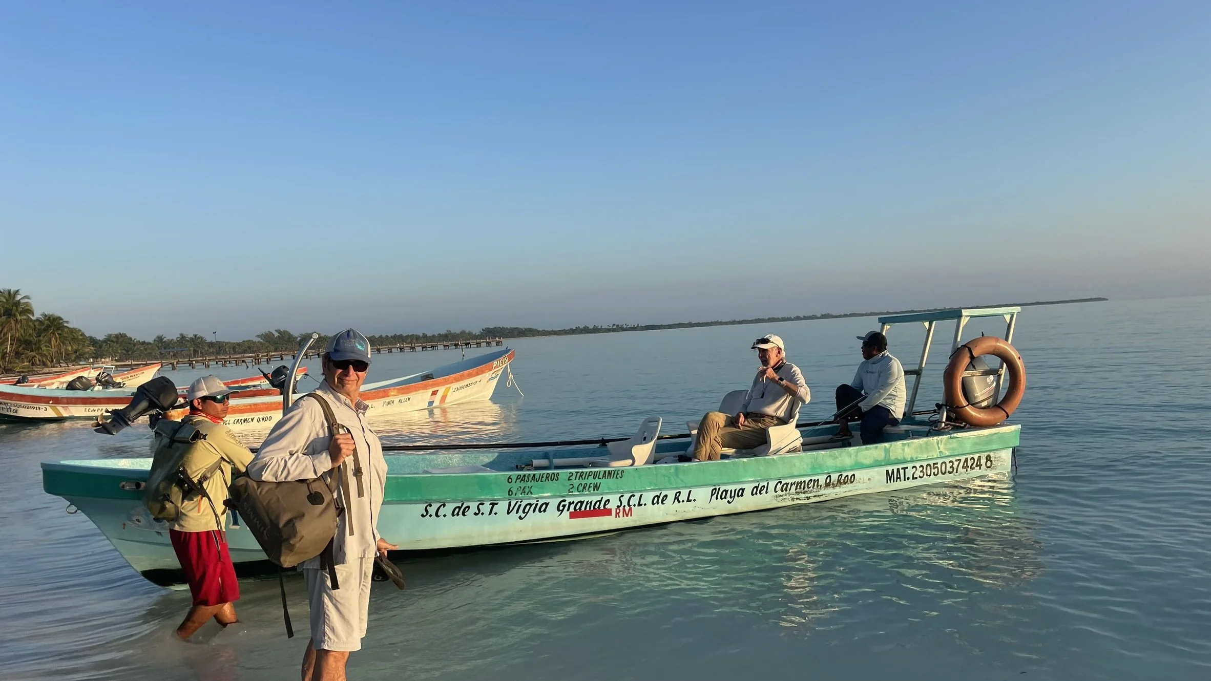Group of four men with backpacks boarding a green boat on calm water, with palm trees and a pier in the background.