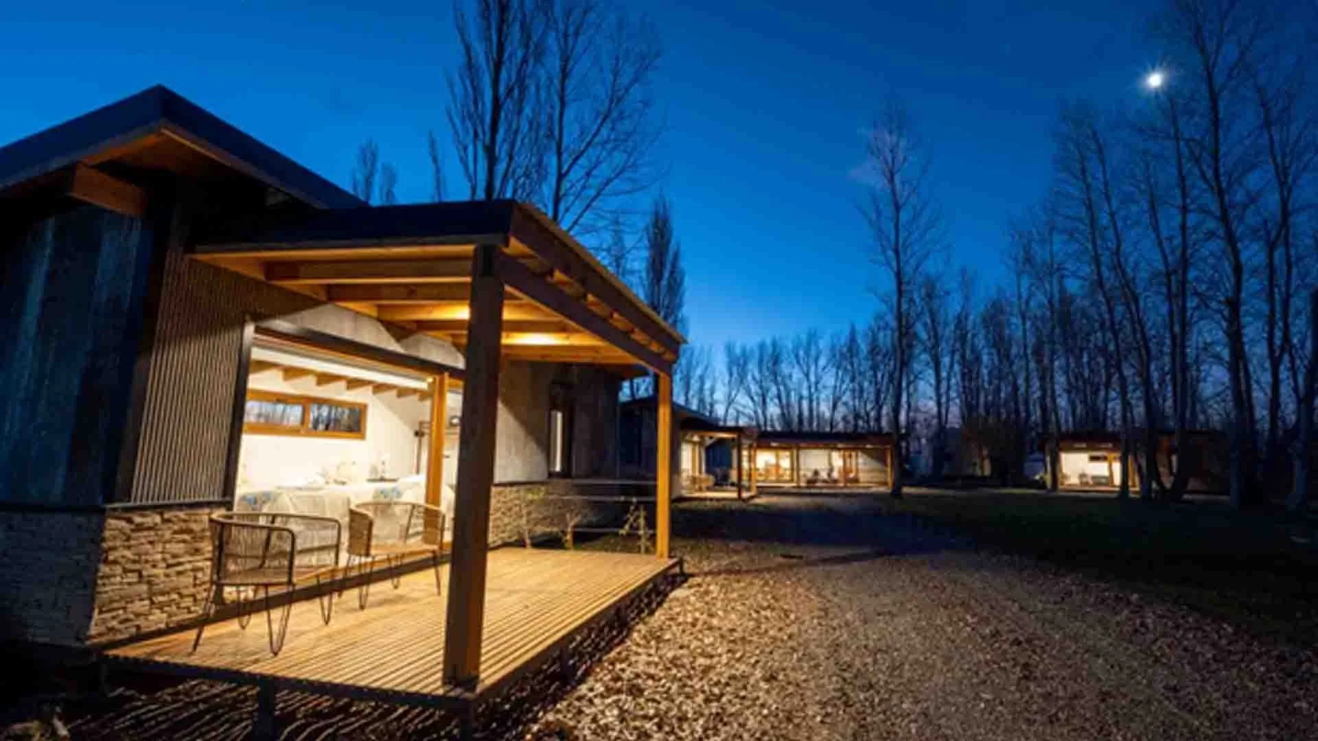 A row of modern cabins with large glass windows and wooden decks, illuminated at dusk, surrounded by leafless trees under a dark blue sky with a visible moon.