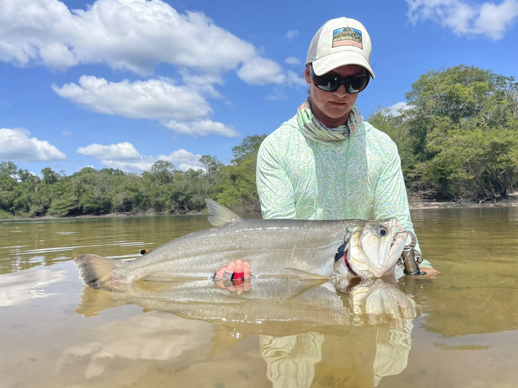 Angler wearing sunglasses, patterned cap, and light green hoodie holding a large Payara (vampire fish) in shallow waters of the Agua Boa River with trees and cloudy sky in Brazil