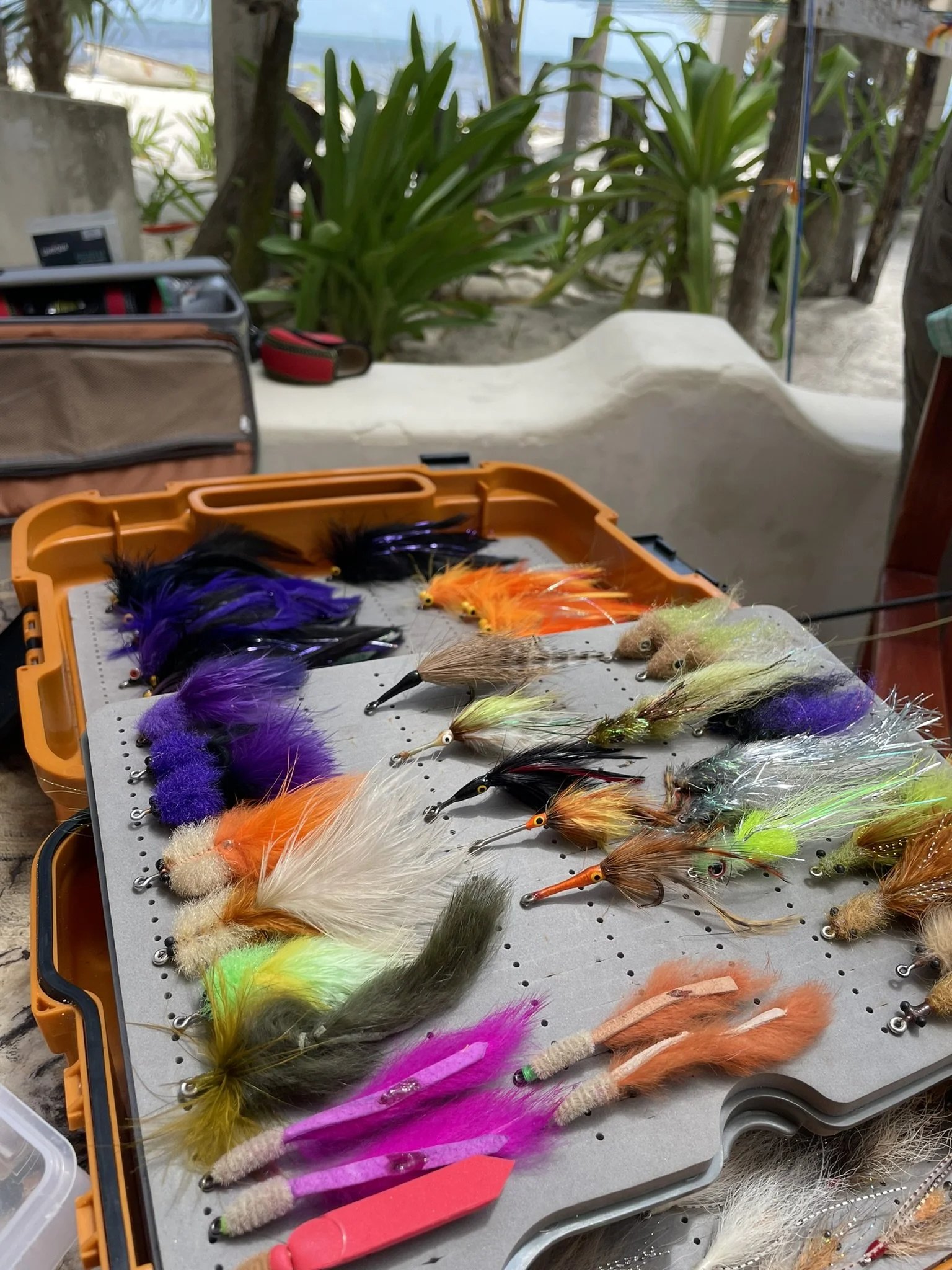 A collection of colorful fishing lures with feathers and fur, arranged on a foam pad inside an orange tackle box on a table outdoors.