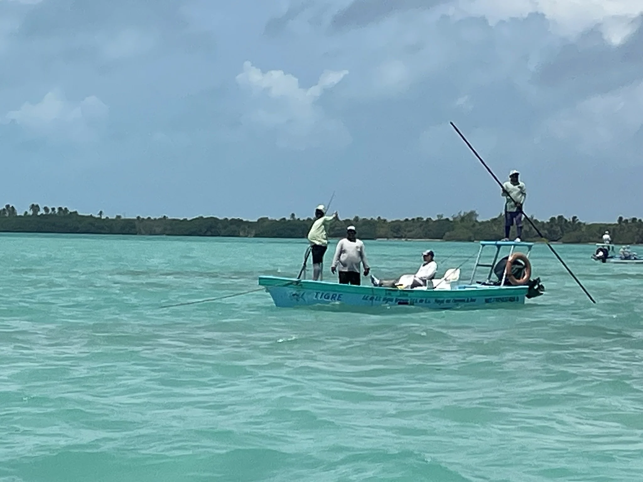 A group of five people on a small blue boat in a turquoise body of water, with one person standing on the front and others seated or standing on the boat, some holding fishing poles, under a cloudy sky.