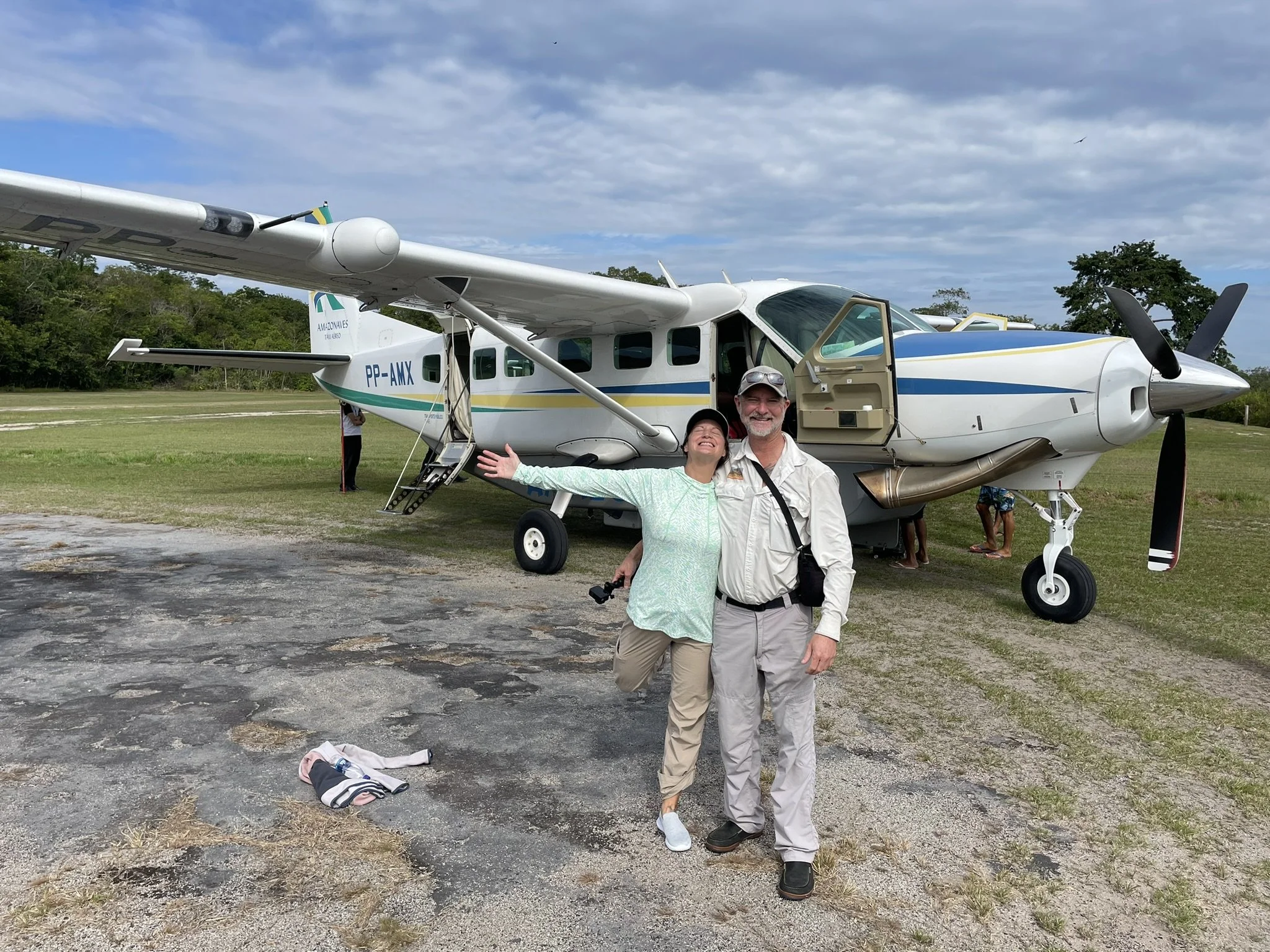 Two people are smiling and happy, standing in front of a small white aircraft with blue and yellow stripes. The woman is gesturing energetically with her arms wide open, and the man is smiling with his arm around her. The scene appears to be at an ai