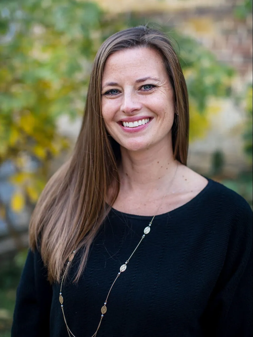 A woman with long brown hair smiling outdoors, wearing a black top and a long necklace.