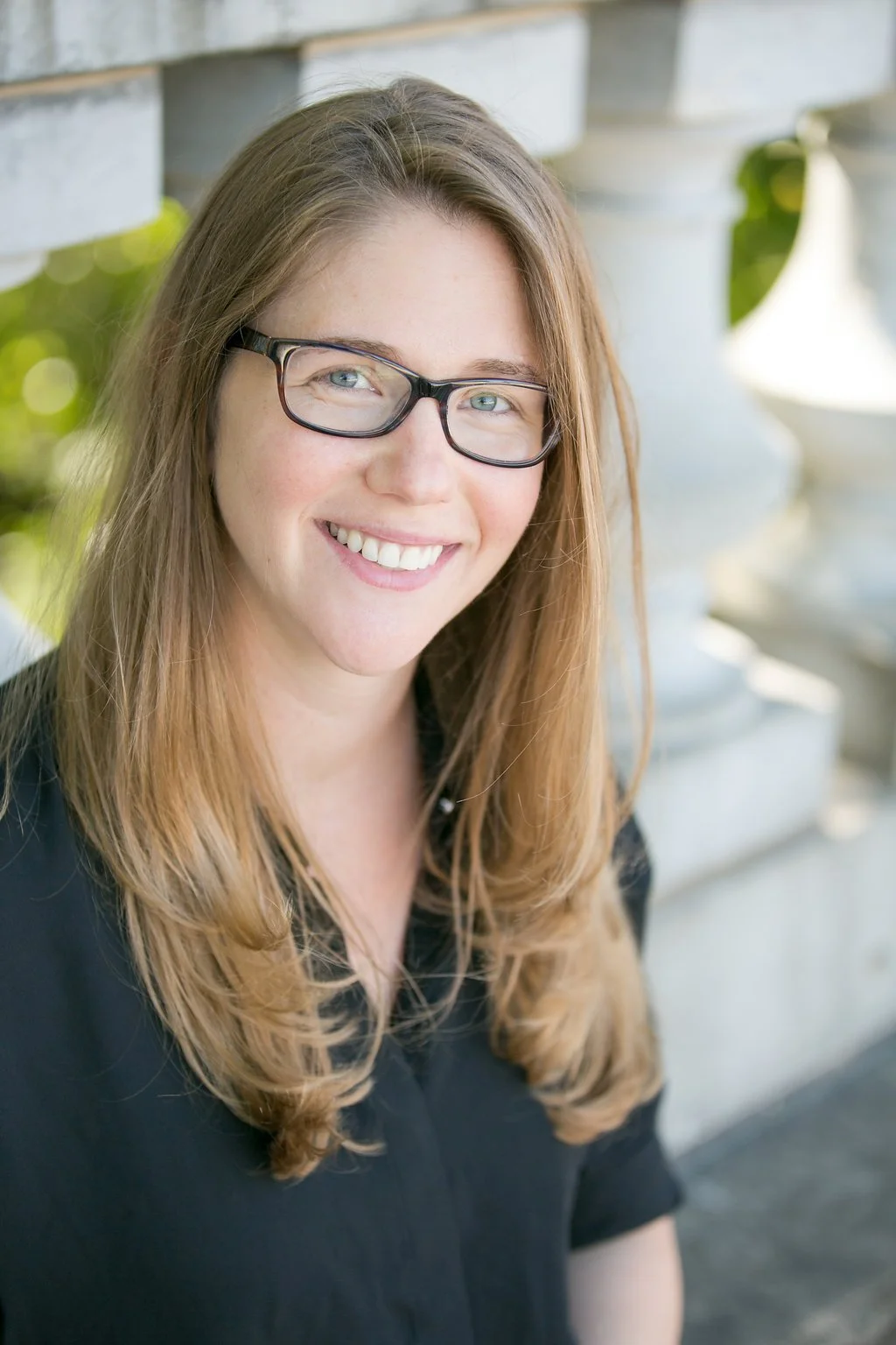 A woman with long, wavy, light brown hair, glasses, and a black top smiling outdoors near white columns and greenery.