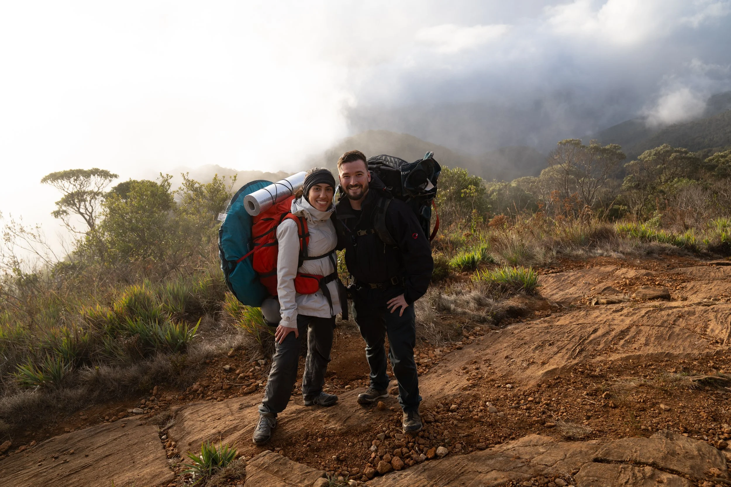 Two hikers, Basil Minder and Juila A. Pinna with backpacks smiling on a rocky trail with mist and trees in the background while on pico do bandeira Brazil