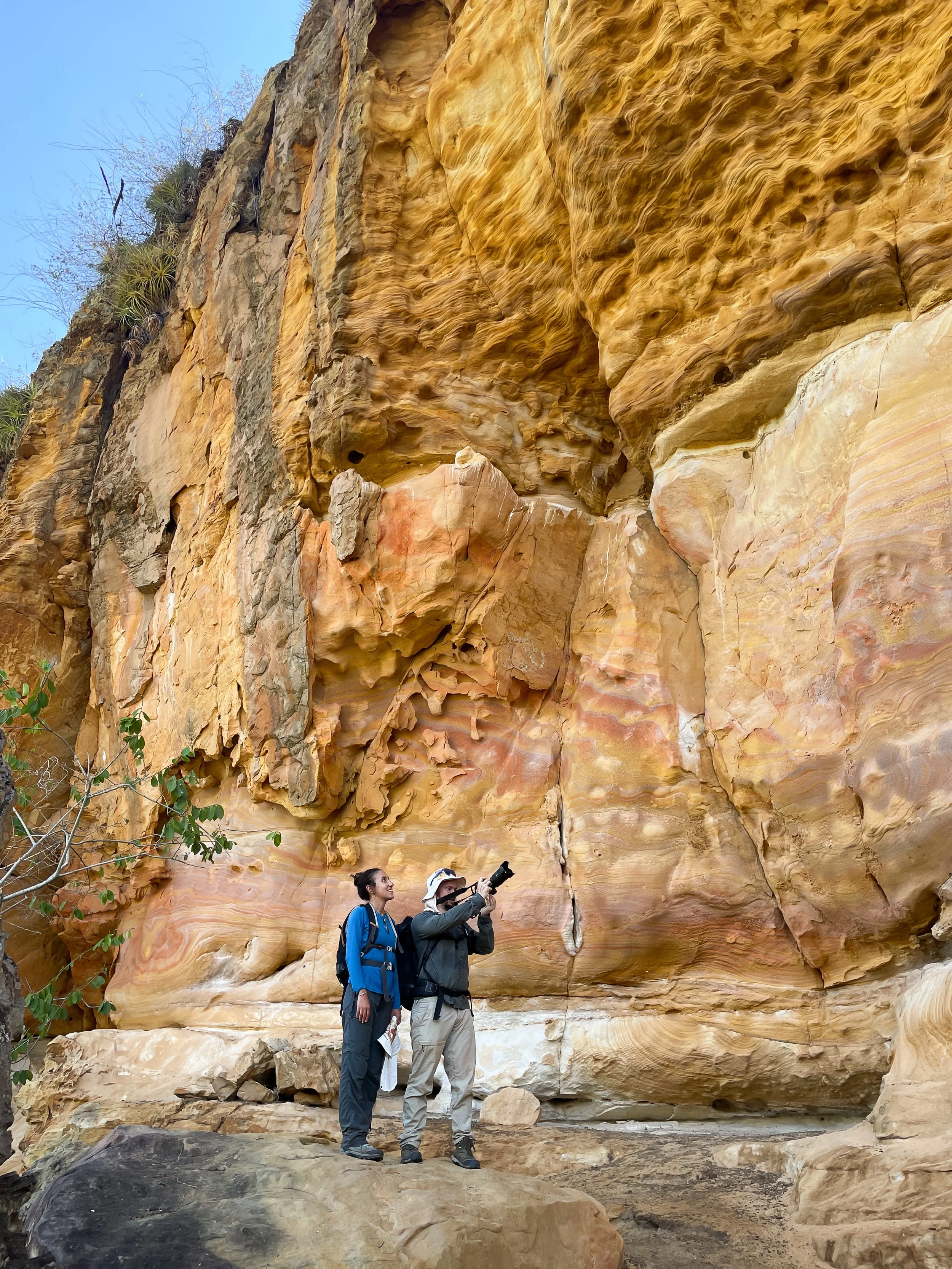 Two people, Basil Minder and Juila A. Pinna with backpacks, one holding a camera, stand in front of a large, colorful rock wall with layered formations. Sparse vegetation is visible at the top of the cliff, in the caatinga.