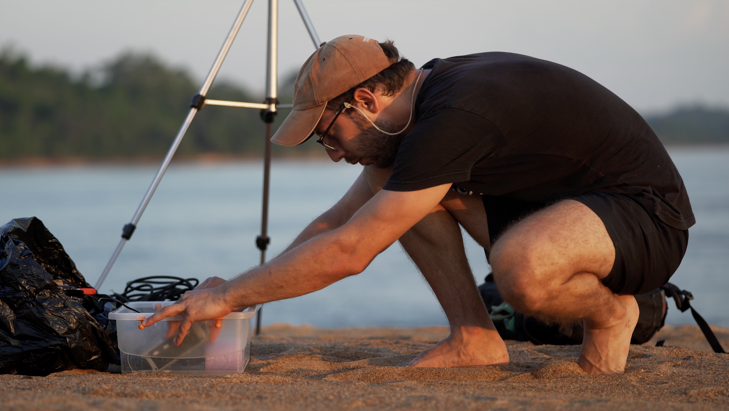 Person in a black shirt and cap crouches on a sandy beach, handling equipment in a plastic container, with a tripod nearby and water in the background.
