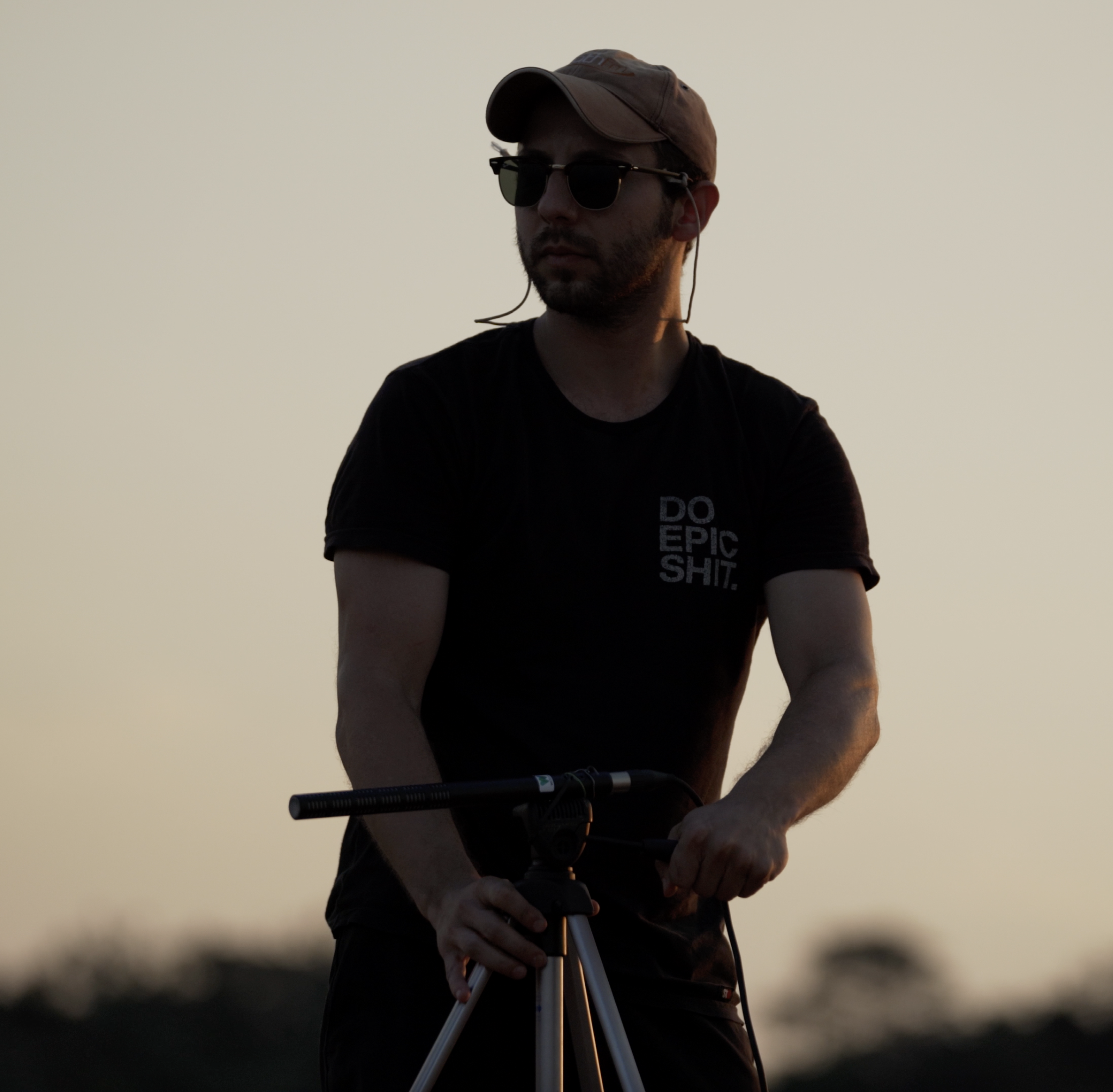 Silhouette of a man operating a tripod during sunset, wearing sunglasses, a cap, and a shirt with text.