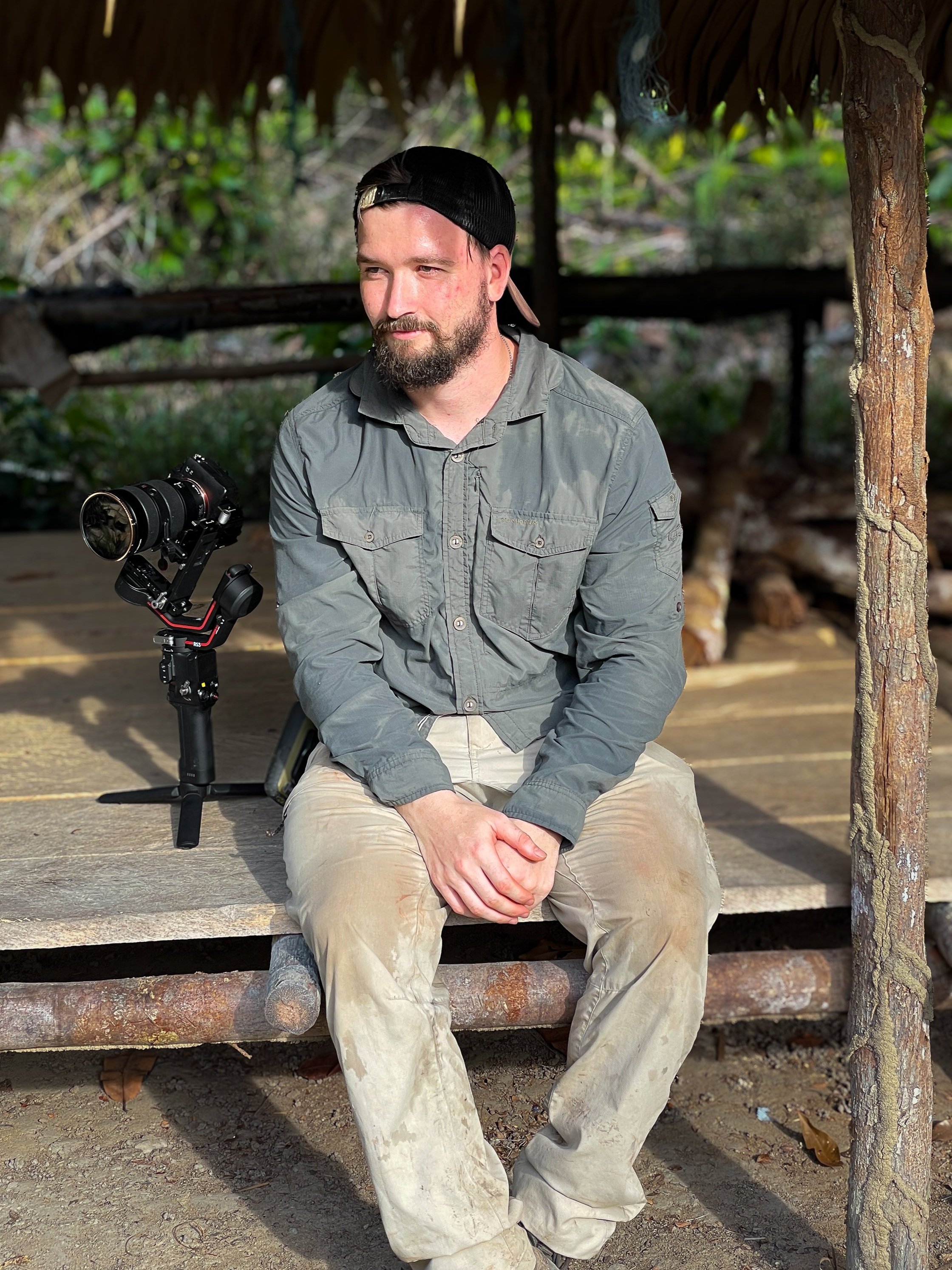 A man wearing outdoor clothing and a backward cap sits on a wooden platform with a camera mounted on a stabilizer beside him. The background is a natural, wooded area. Basil Minder