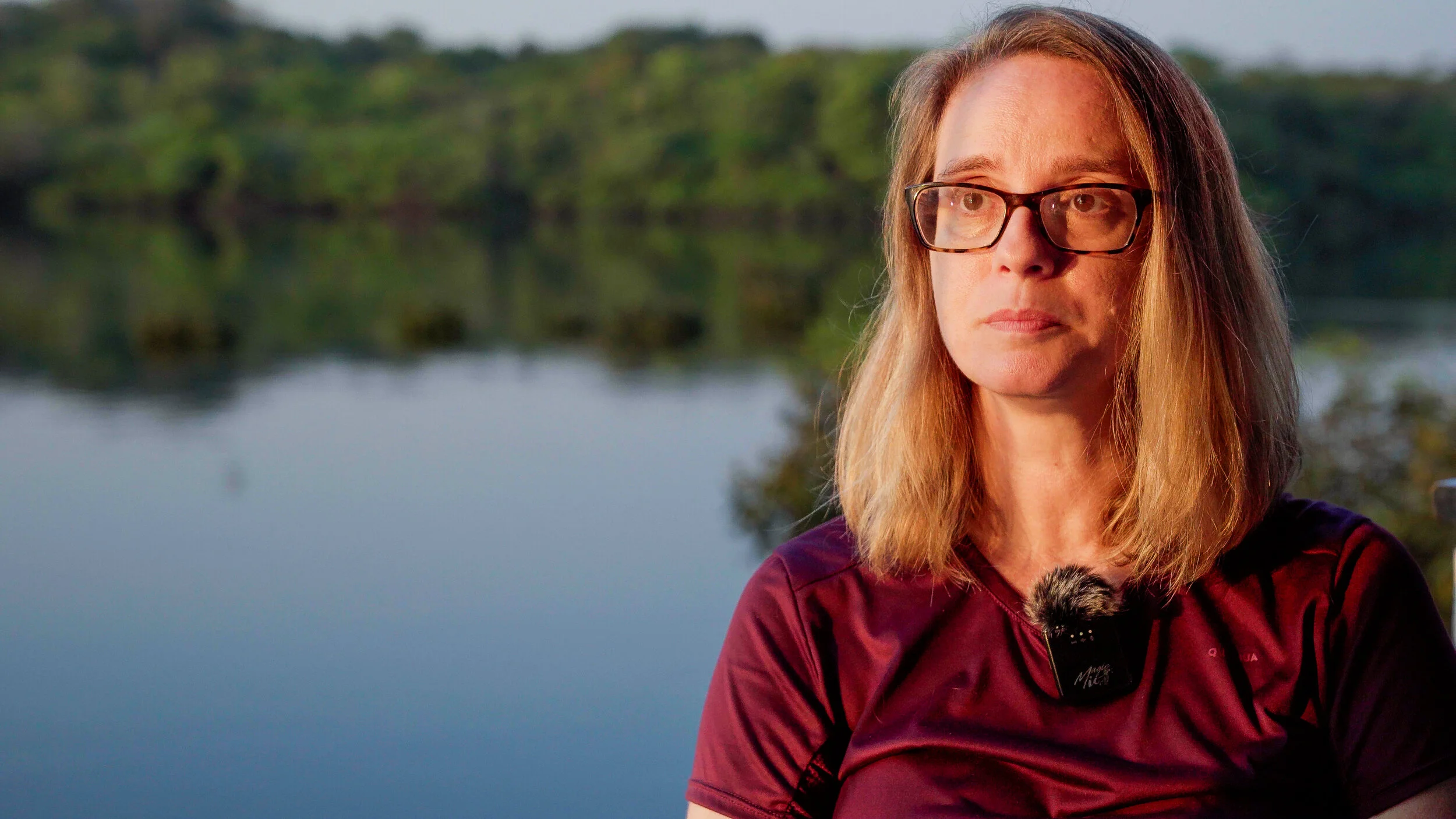 A woman with glasses and a purple shirt standing outdoors near a body of water with trees in the background.