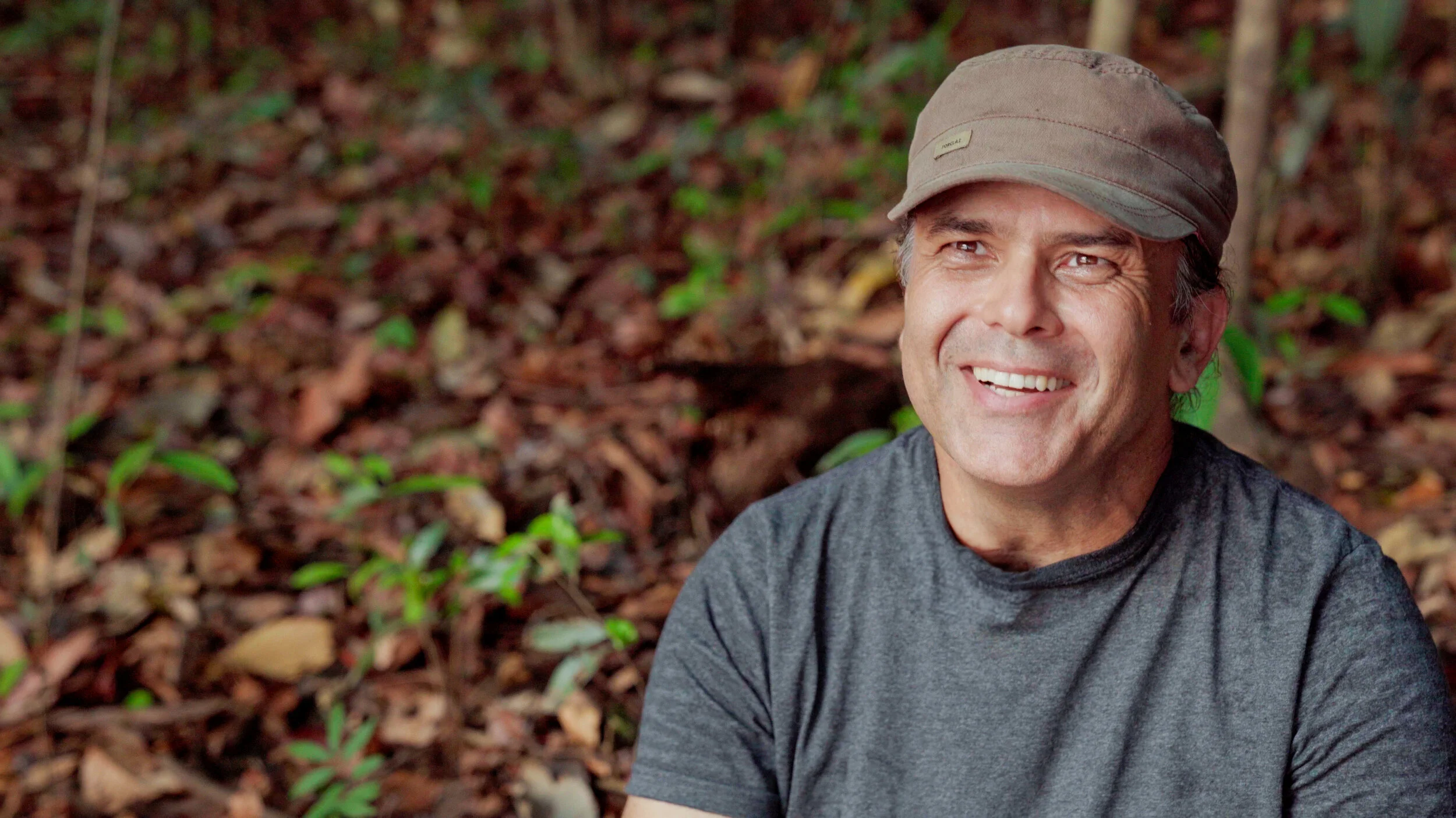 Smiling man wearing a gray shirt and hat in a forest setting.